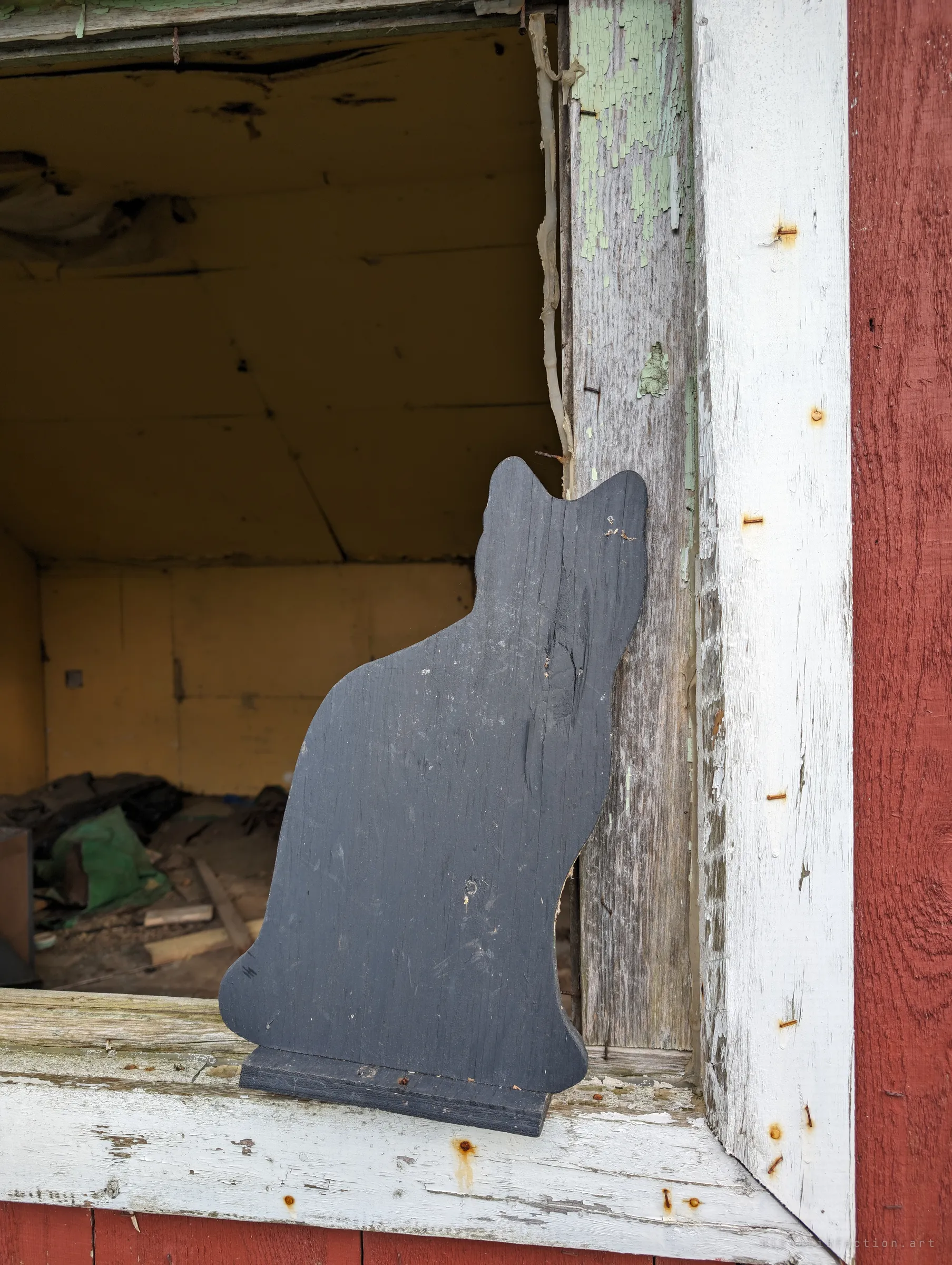 A black wooden cat cut-out sits on the peeling sill of the shed's empty window frame, the wrecked yellow interior visible behind it. A small decorative touch that outlasted everything else.