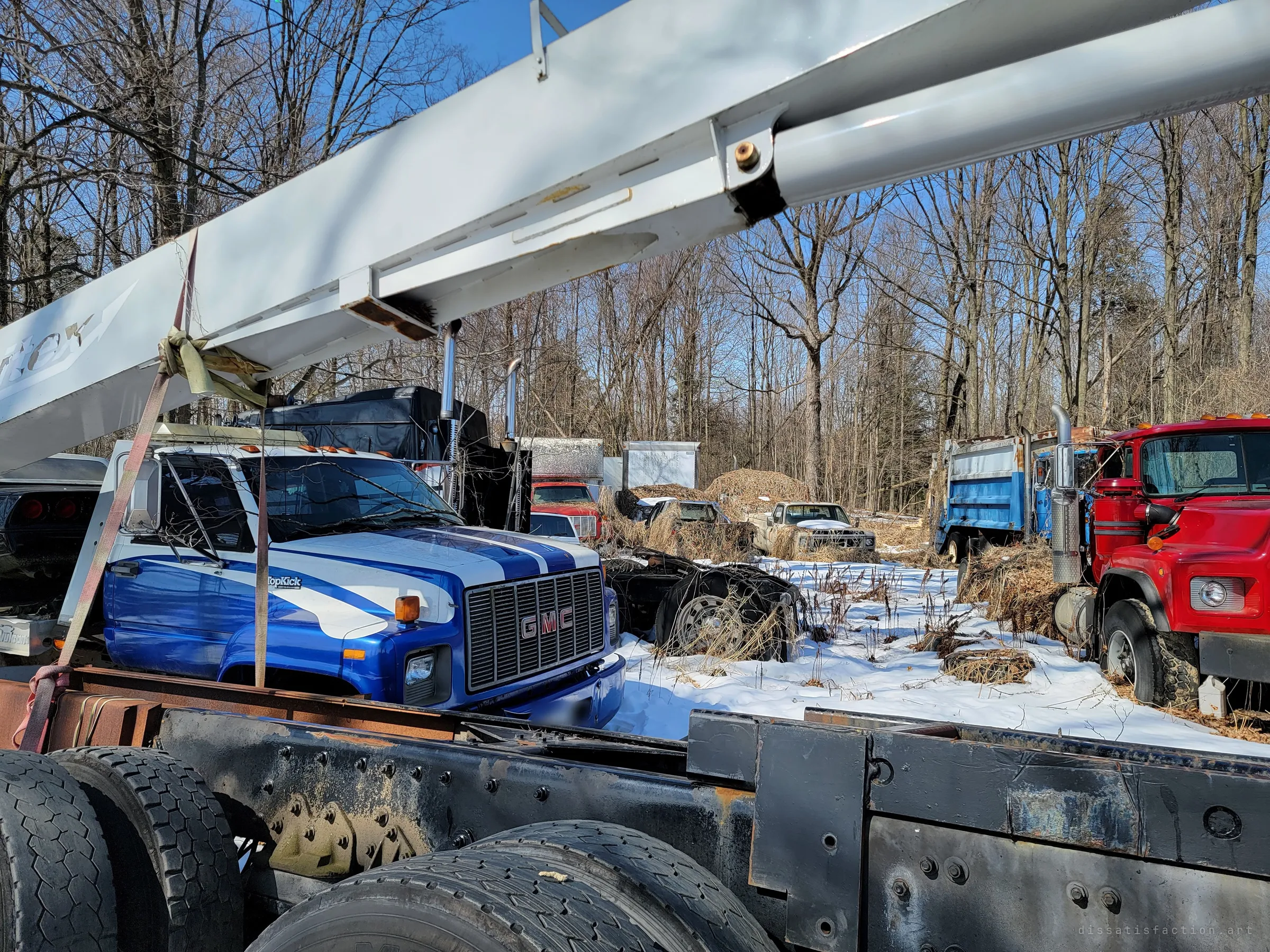 Looking out from under a white crane boom across a snowy yard of parked trucks, a blue-and-white striped cab-over truck in front and a red dump truck to the right. Dozens of work vehicles sit abandoned among bare winter trees.