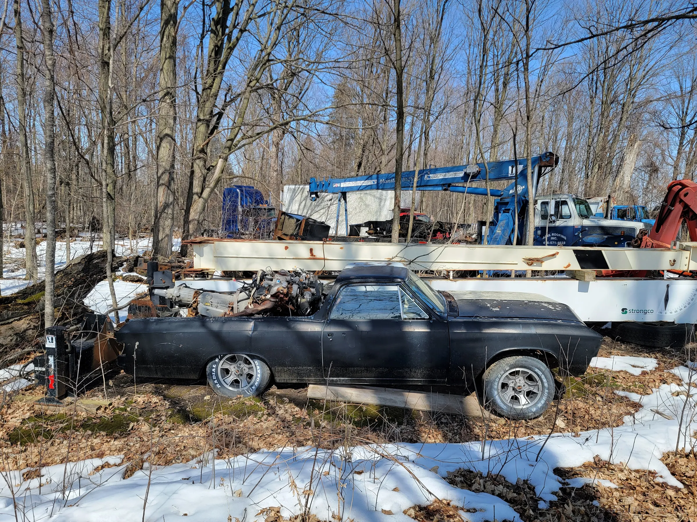 A black classic car-based pickup with its engine and transmission dumped in the bed, parked in the woods beside a long white crane boom and a blue knuckle crane. Snow and leaf litter cover the ground.