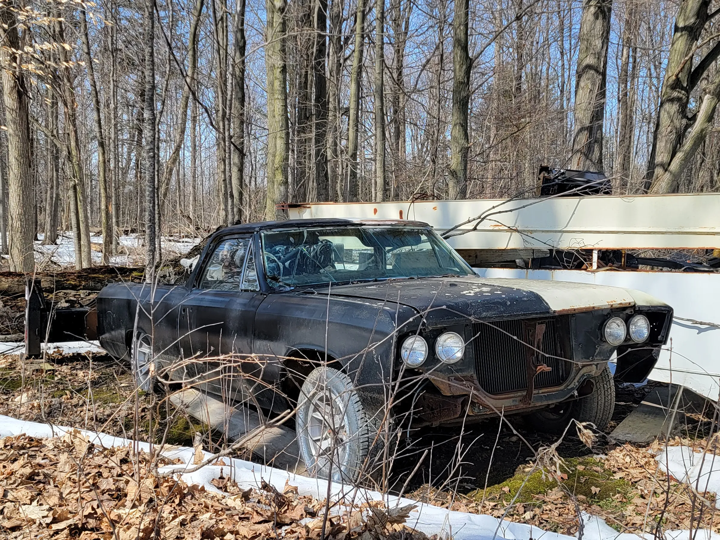 Three-quarter view of the same black pickup with a bare radiator and quad headlights, grille and bumper missing. It rests on boards among dead leaves next to the white boom.