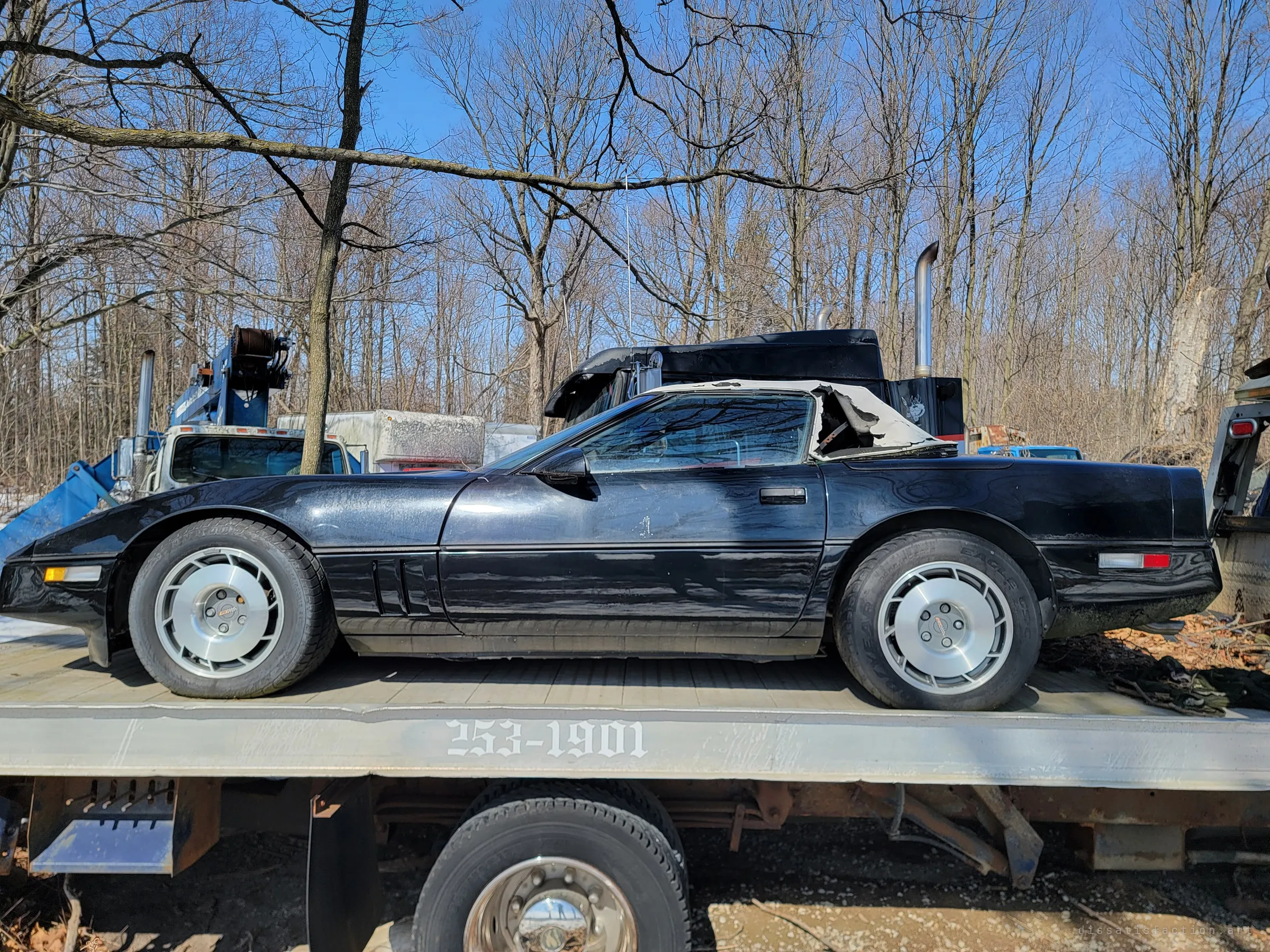Side profile of a black convertible sports car with a torn white soft top, strapped on the flatbed of a tow truck. A heavy tractor unit and blue crane truck stand behind it in the bare woods.