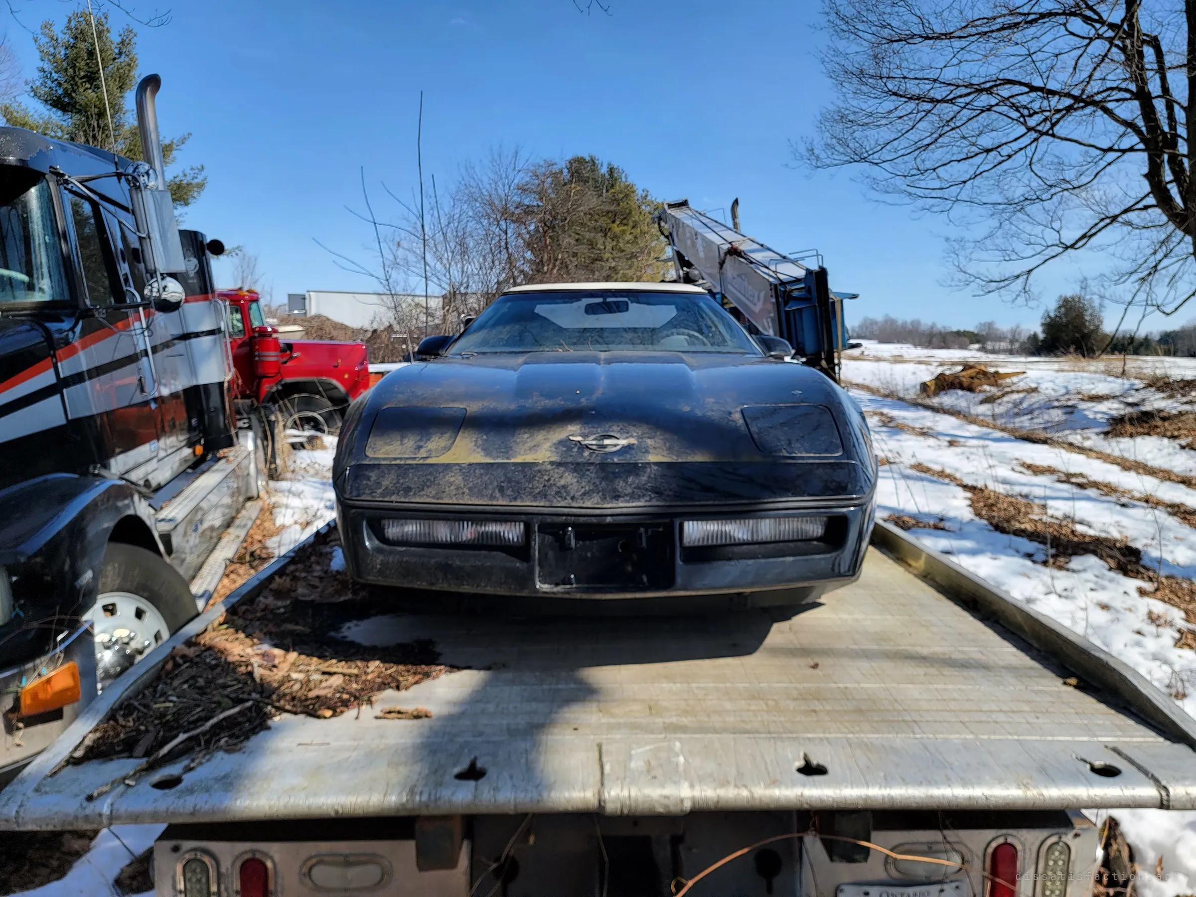 Low, wide view down the flatbed at the sports car's dusty nose, a tractor truck on the left and open snowy fields on the right. The yard spills out of the woods here.