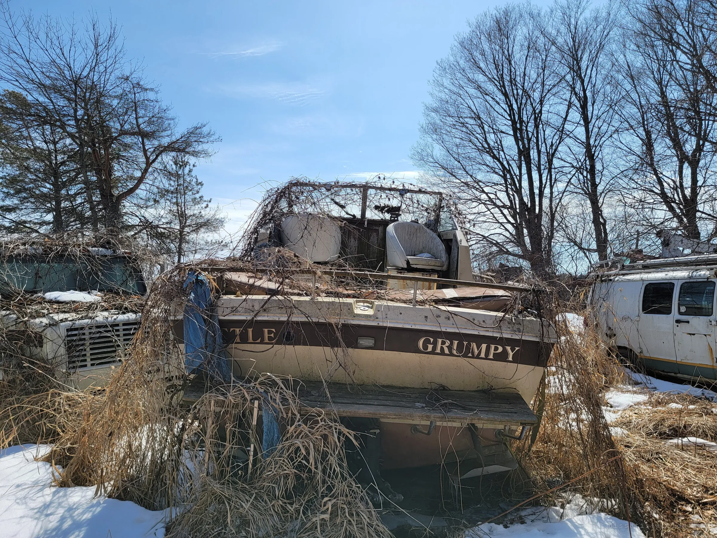 A cabin cruiser sits beached in dry grass and snow, its windshield frame wrapped in dead vines, between an old truck cab and a rusting utility van. A boat stranded far from any water sets the tone of the yard.