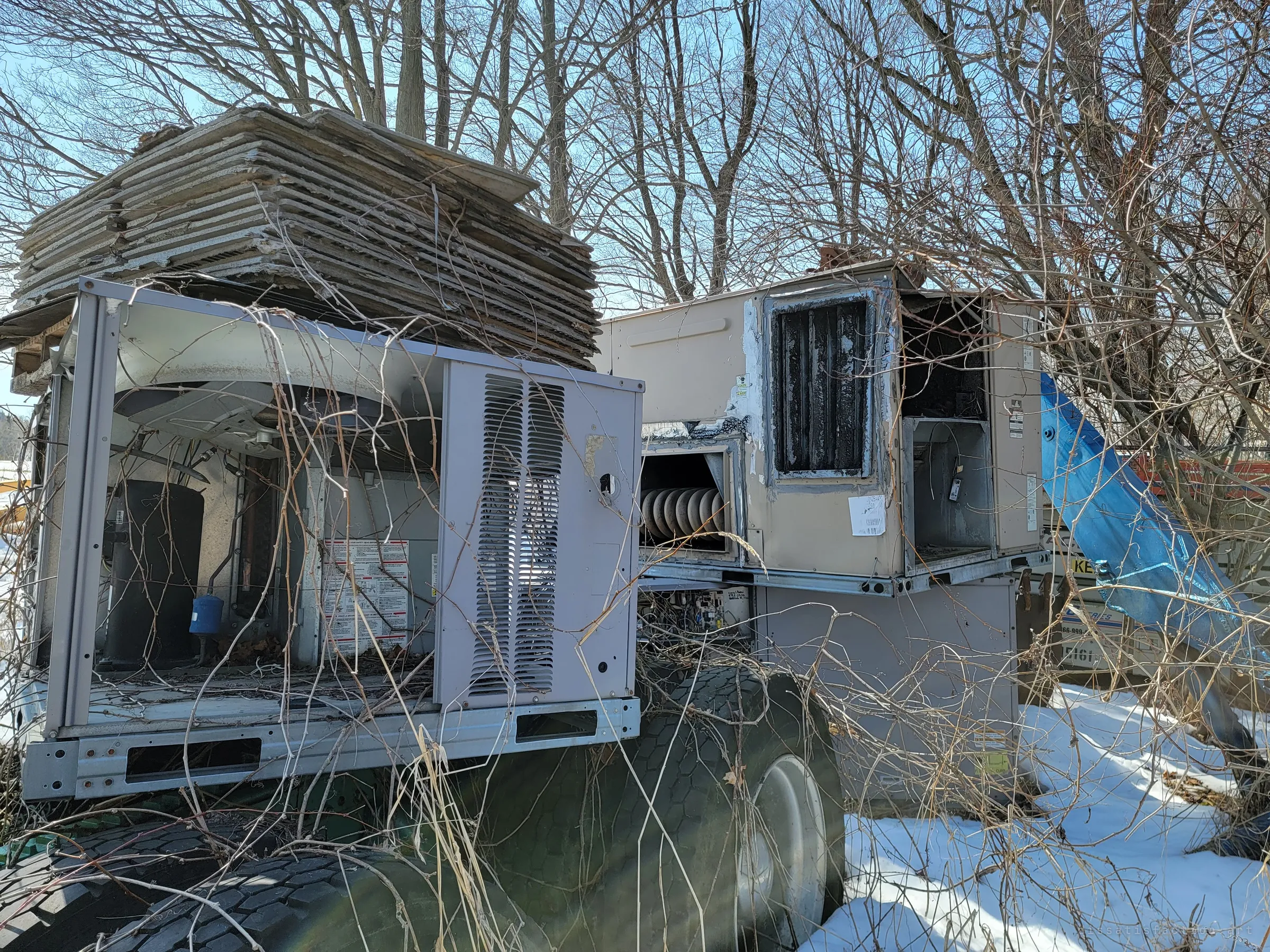 Two gutted rooftop HVAC units sit on a trailer under a stack of weathered panels, their fans and coils exposed and tangled in bare vines. Even the machinery that once cooled buildings has been left here to rot.