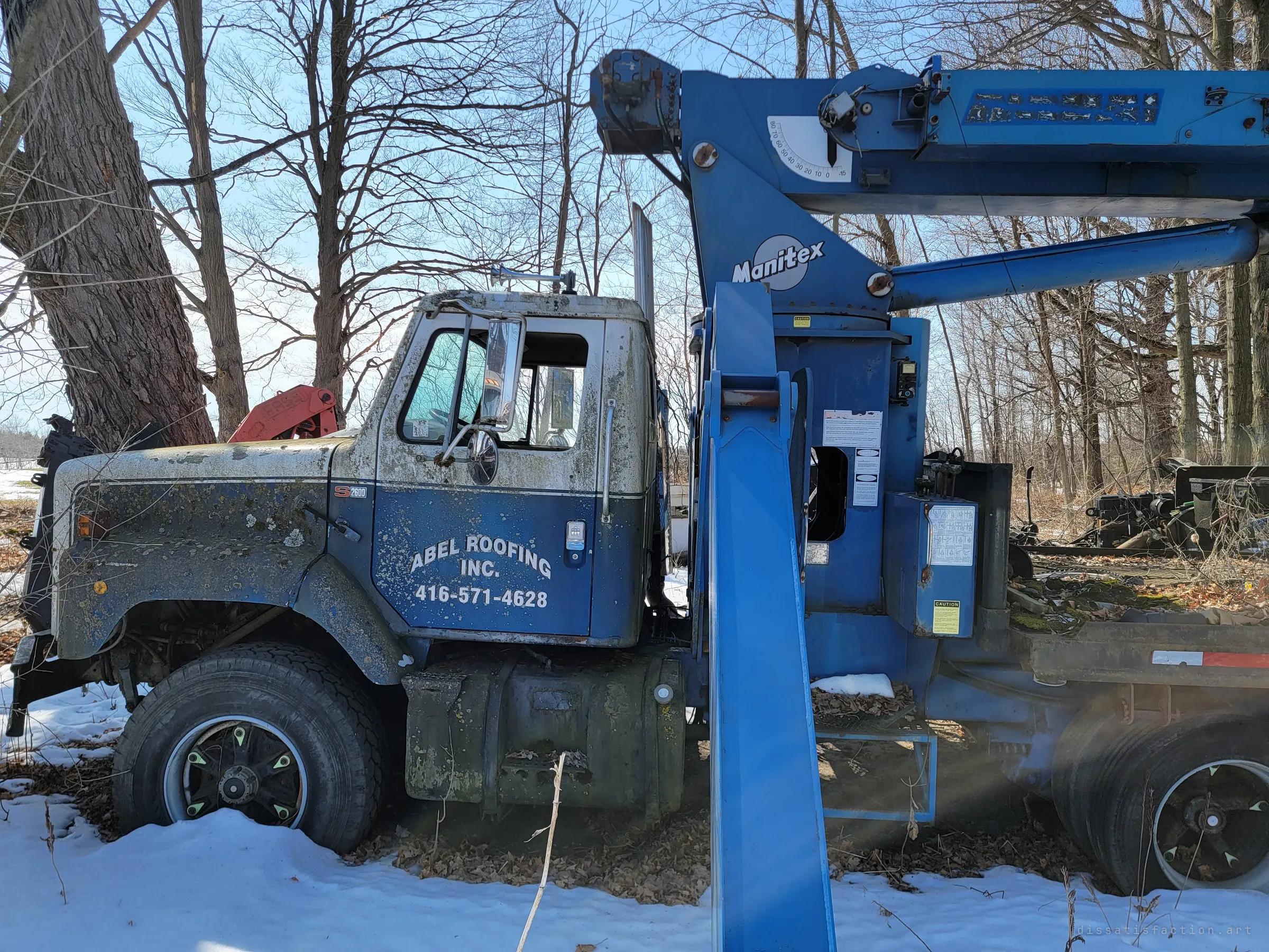 A lichen-covered boom truck with a blue crane mast stands in snow among tall bare trees, its cab paint flaking and mottled. The crane arm still points skyward as if the job was simply abandoned mid-shift.