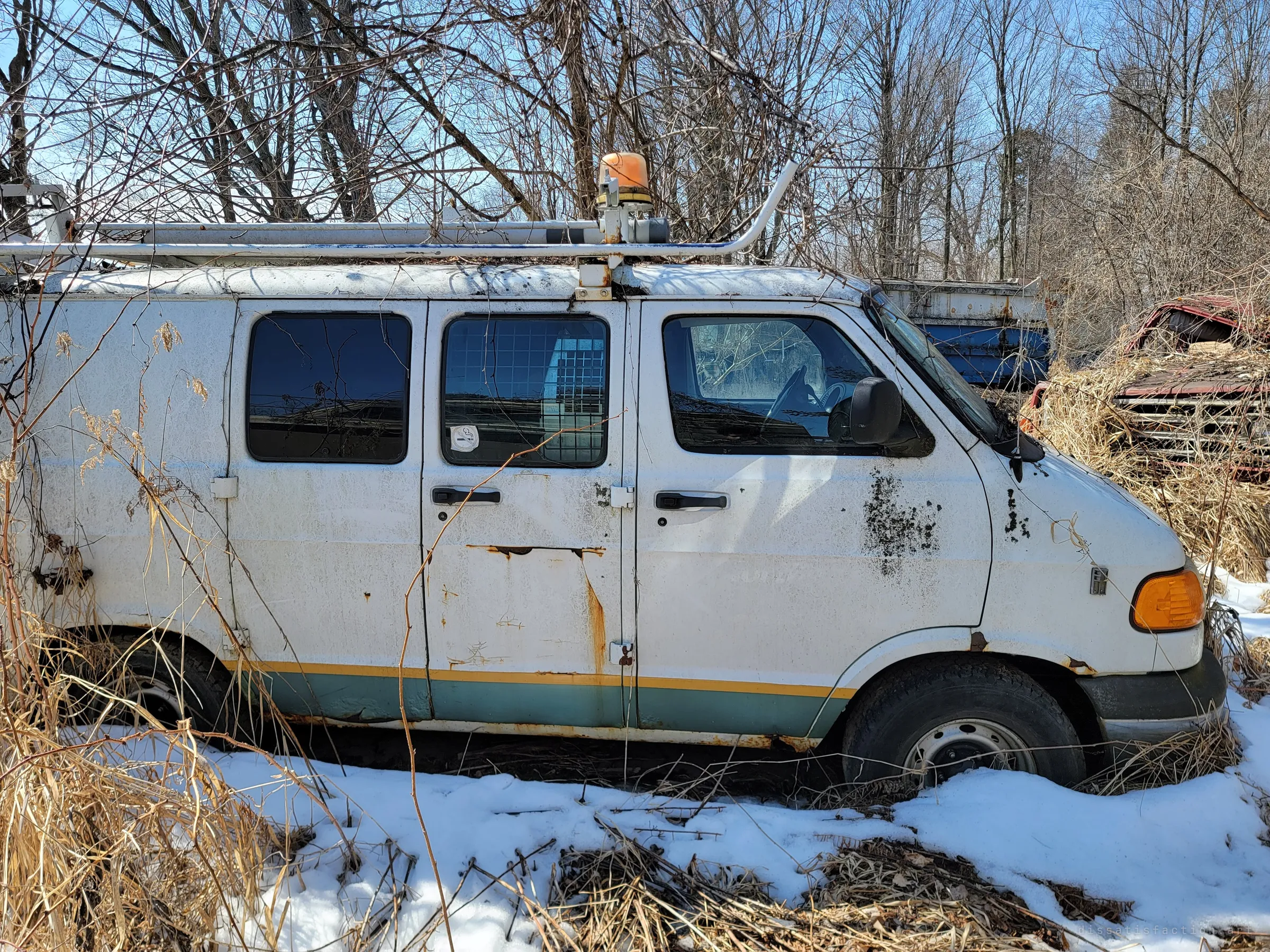 A white work van with a roof rack and amber beacon sinks into snow and dry grass, rust bleeding from its doors and mould spotting its side. The wreck of an old red truck peeks out of the brush behind it.