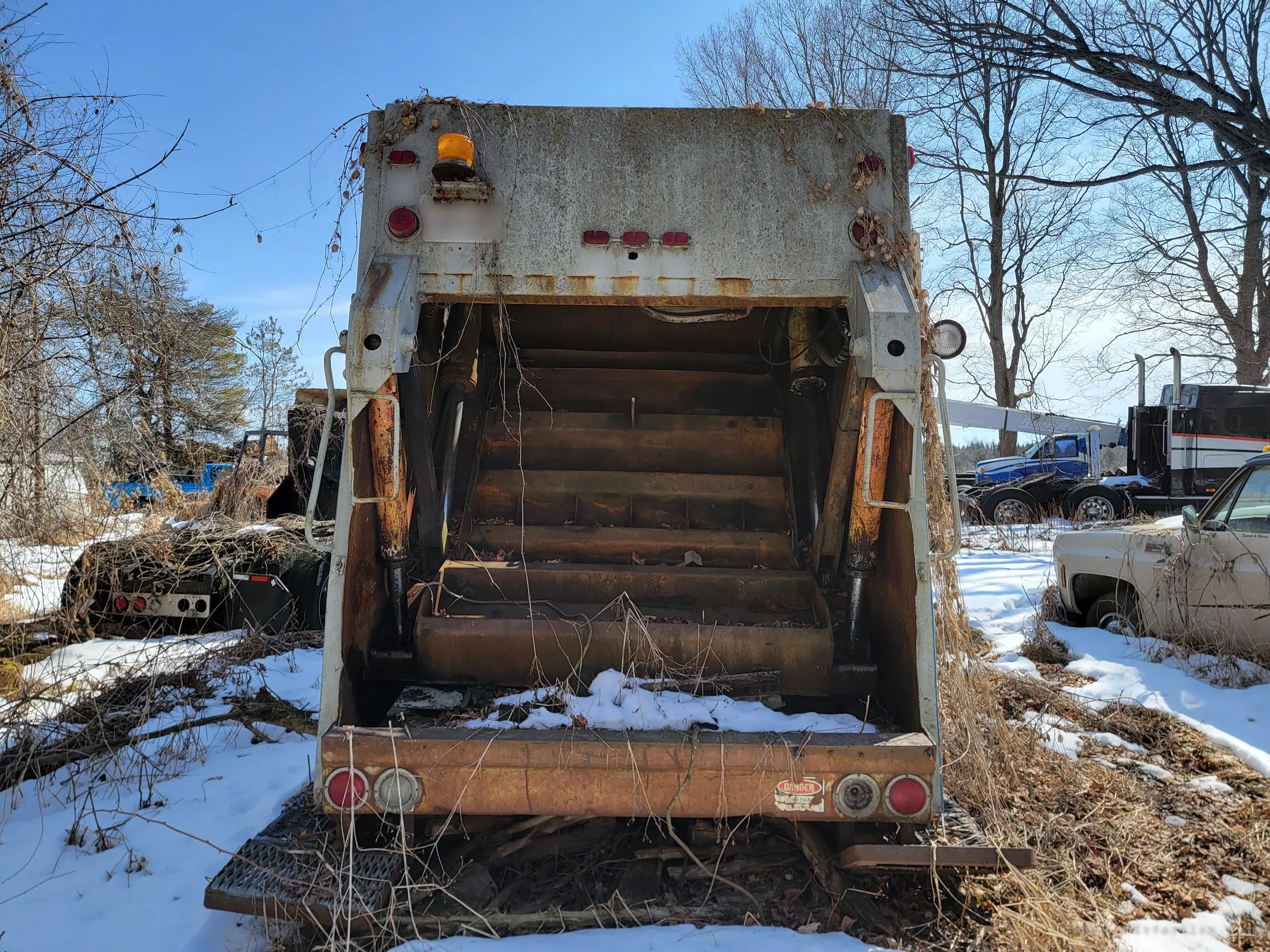 The open rear hopper of a garbage truck, its rusted packer blade and steps dusted with snow, faces the camera head-on. Old pickups and a tractor unit wait in the snow on either side.