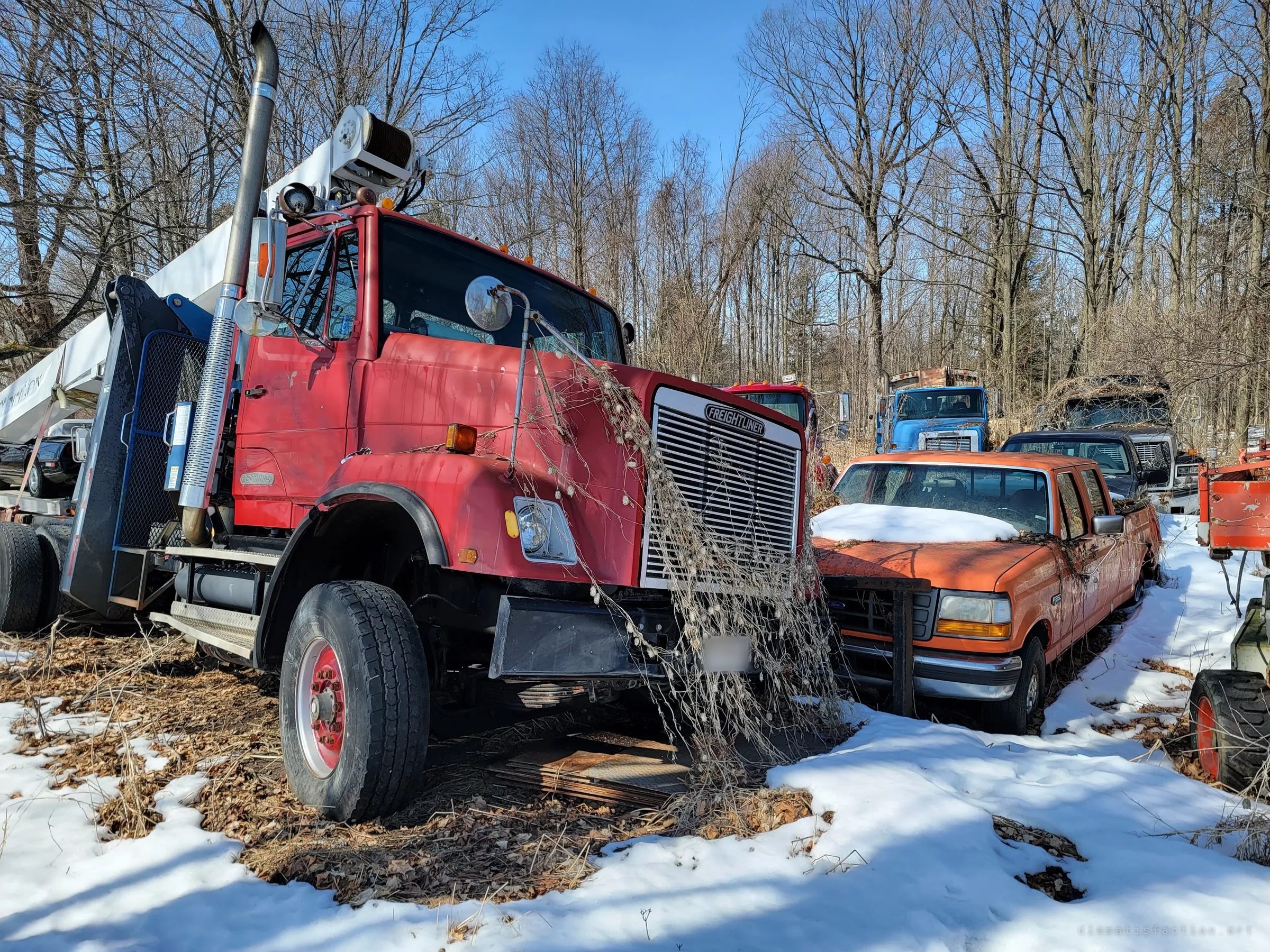 A red heavy truck with a white boom arm stands in the snow, dead vines hanging from its mirror across the grille, next to an orange pickup buried to the windshield in snow. More trucks line up behind them into the woods.