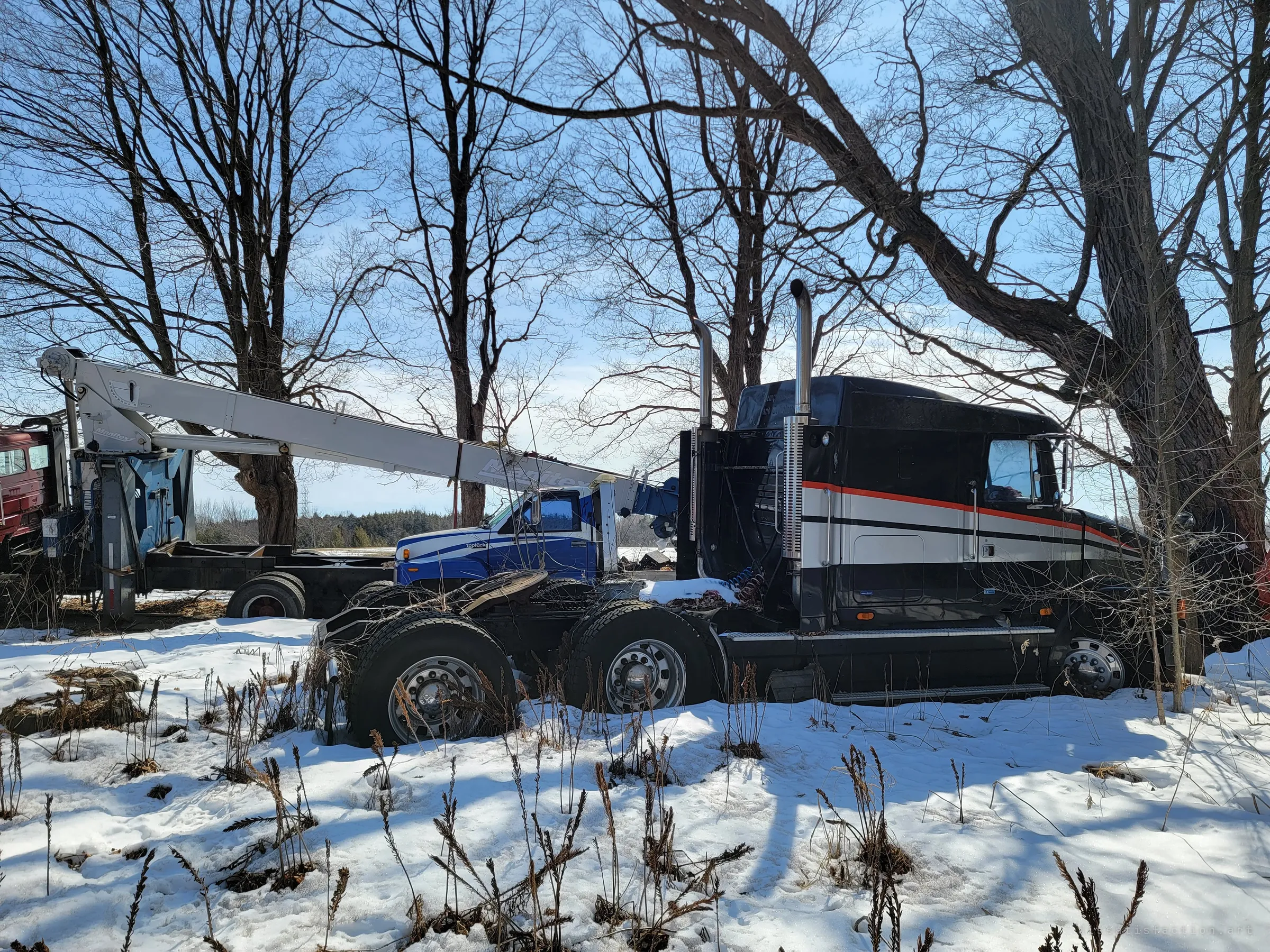 A black long-nose semi tractor with red and white stripes sits in the snow under bare trees, a boom truck's crane stretched out behind it. Heavy trucks parked for good at the edge of a field show the scale of the vehicle graveyard.