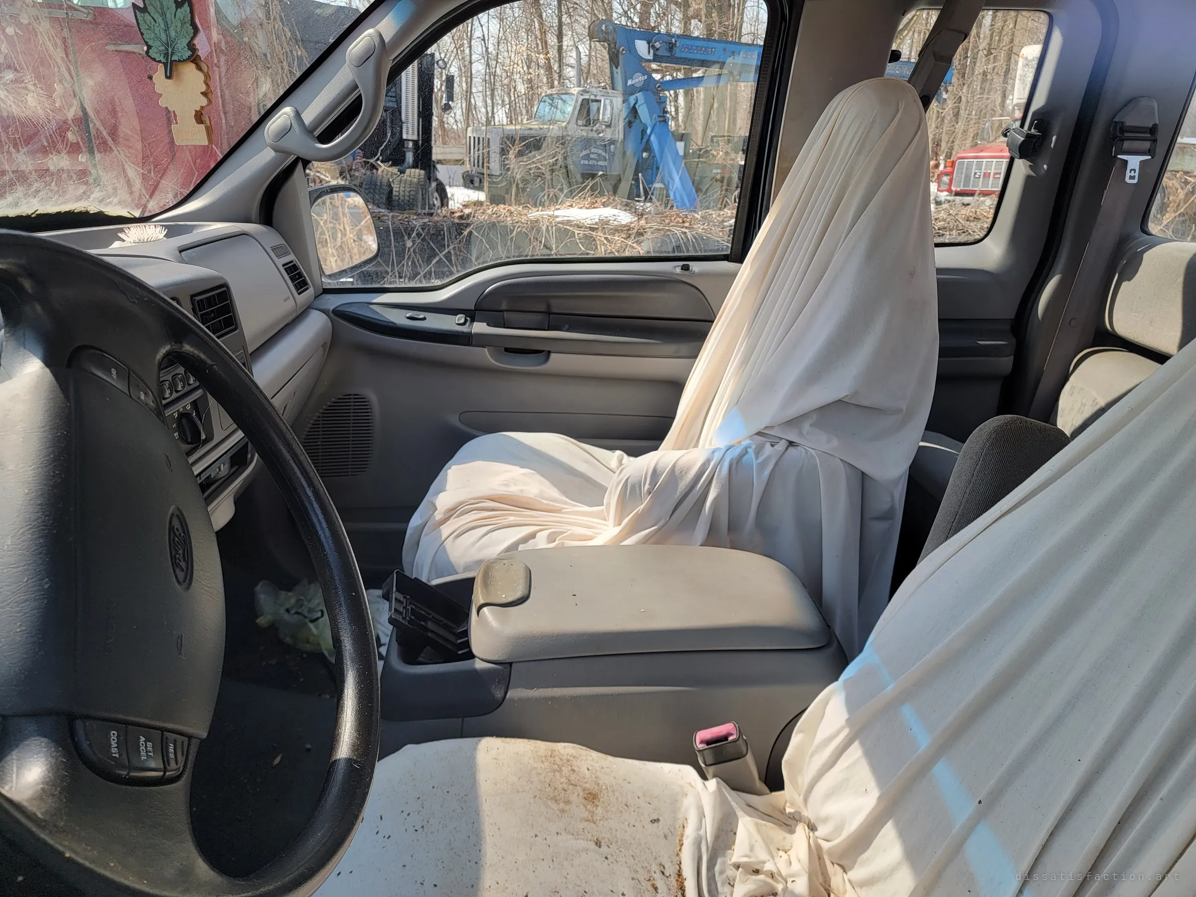 Inside the pickup, both front seats are draped in white sheets like seated ghosts, a tree-shaped air freshener hanging by the windshield. Through the side window a tow truck with a blue crane rots among the brush.