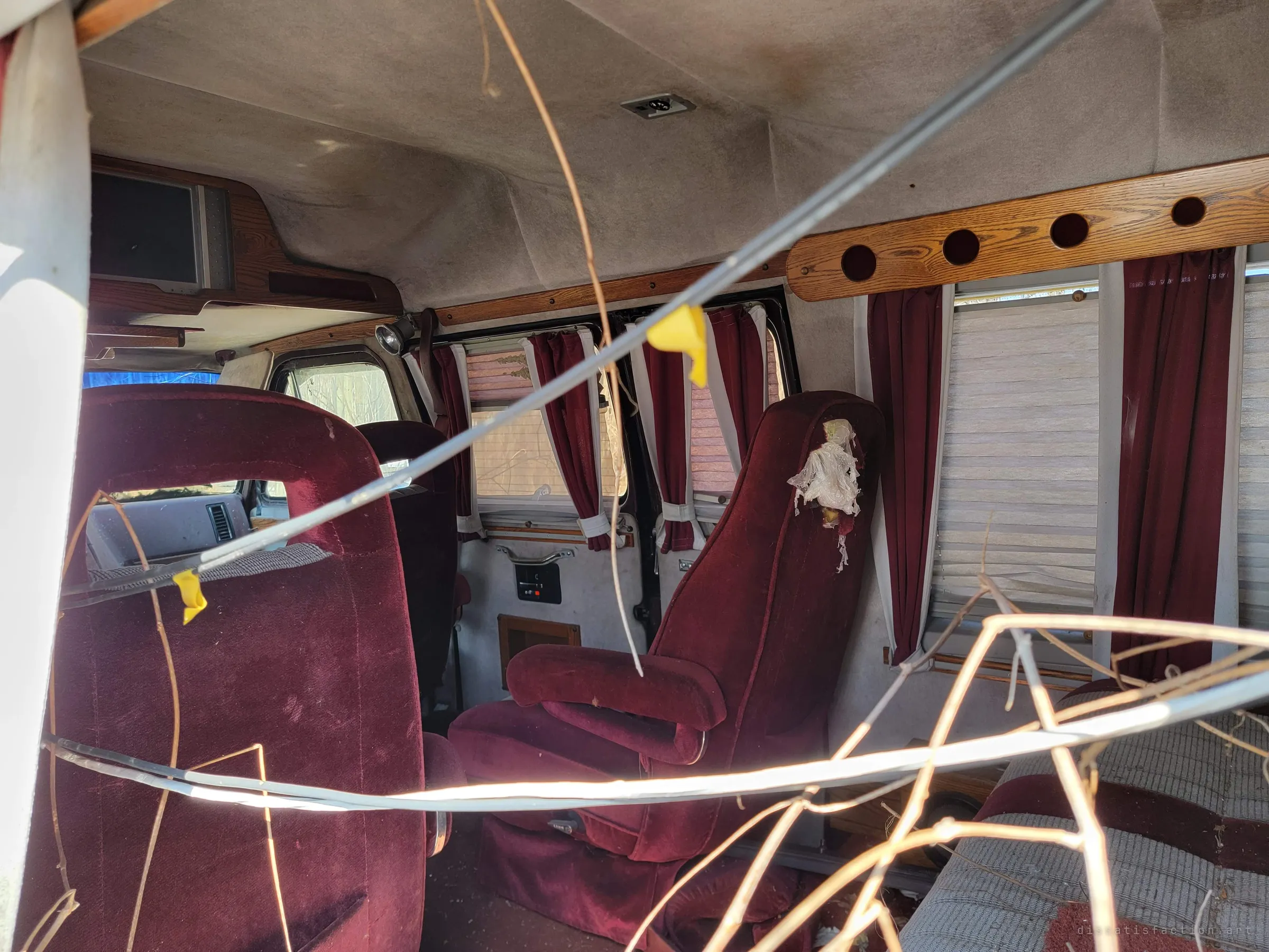 Inside the conversion van, plush red captain's chairs, wood trim and tied-back red curtains sit under a stained headliner, with branches and a loose cable poking in. The dated luxury interior has been left to the weather.