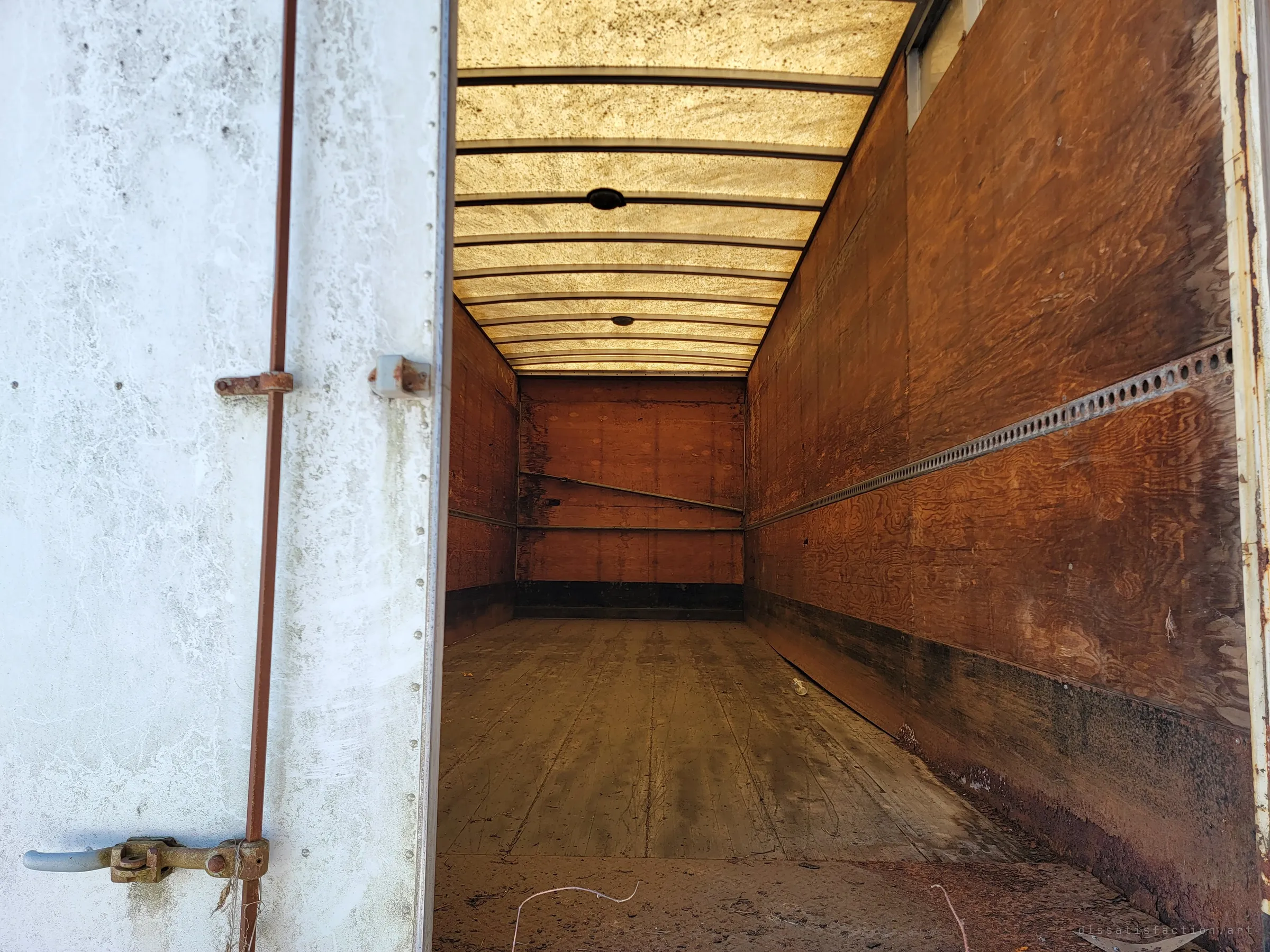 The open door of a box trailer reveals an empty plywood interior lit amber through its translucent roof panels. The long, bare cargo space feels like a tunnel.