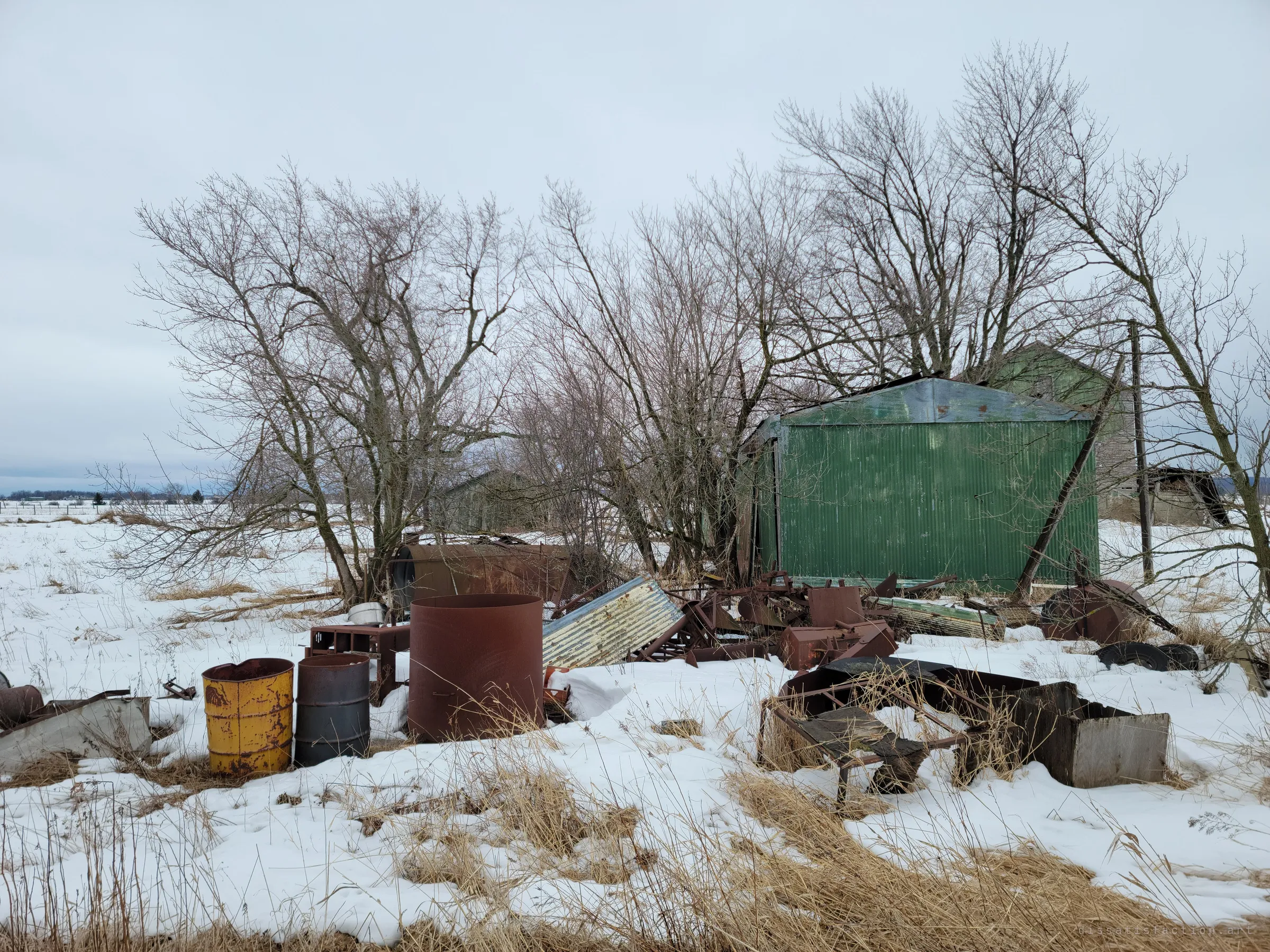 Rusted barrels, scrap metal and farm junk half buried in snow beside a green corrugated shed under bare trees. The yard shows how long the farm has been left to the weather.