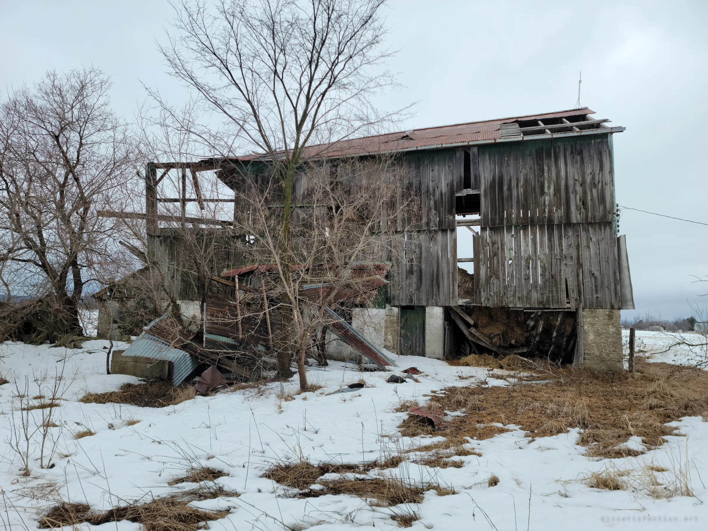 A weathered grey board barn on a concrete block base, its rusted roof peeled open and a lean-to collapsed into scrap beside it. Hay spills out from the ground floor onto the snow.