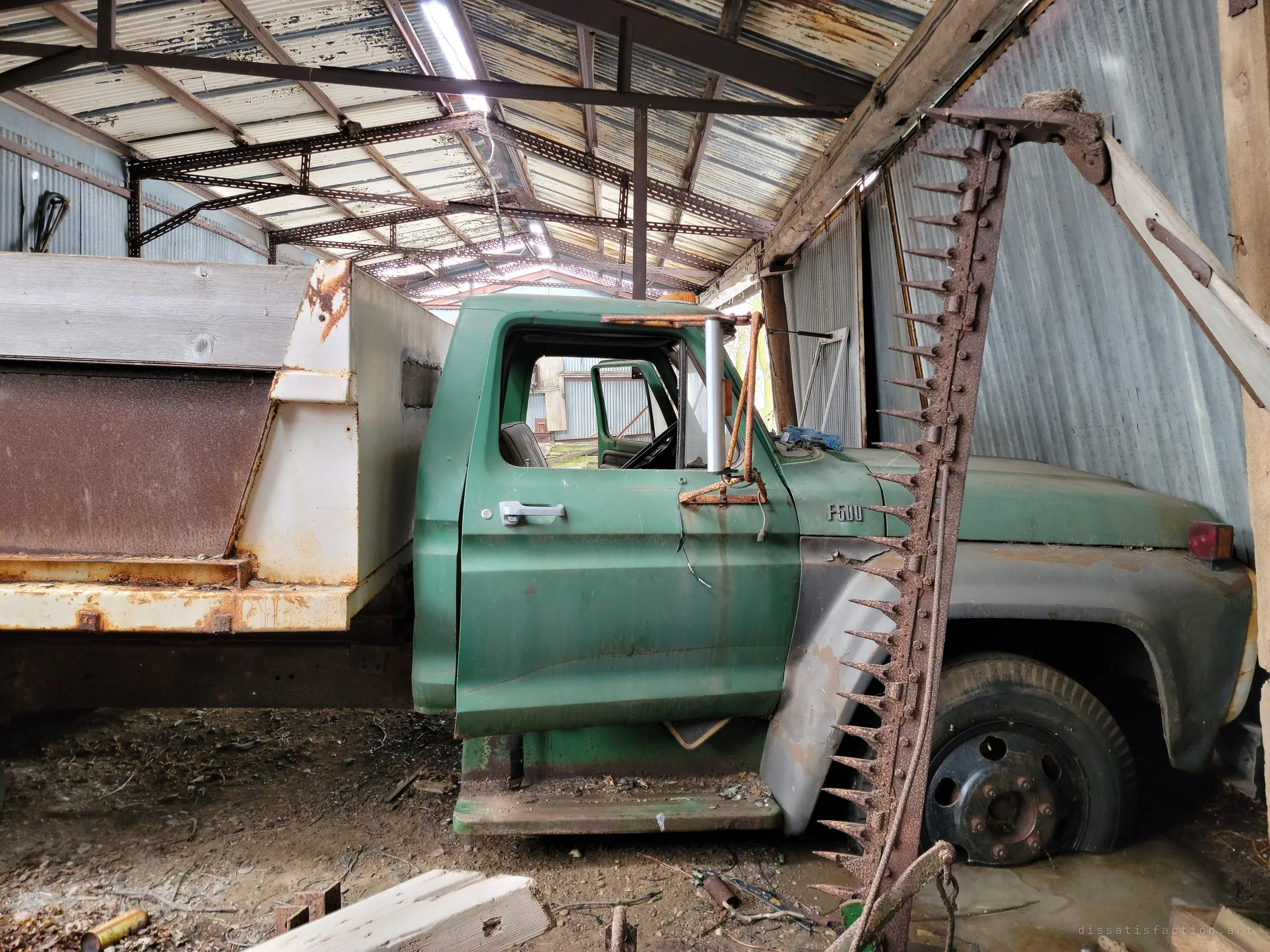 An old green dump truck parked inside a corrugated metal shed, a rusted sickle-bar mower leaning against its fender. Light leaks through gaps in the peeling roof above.
