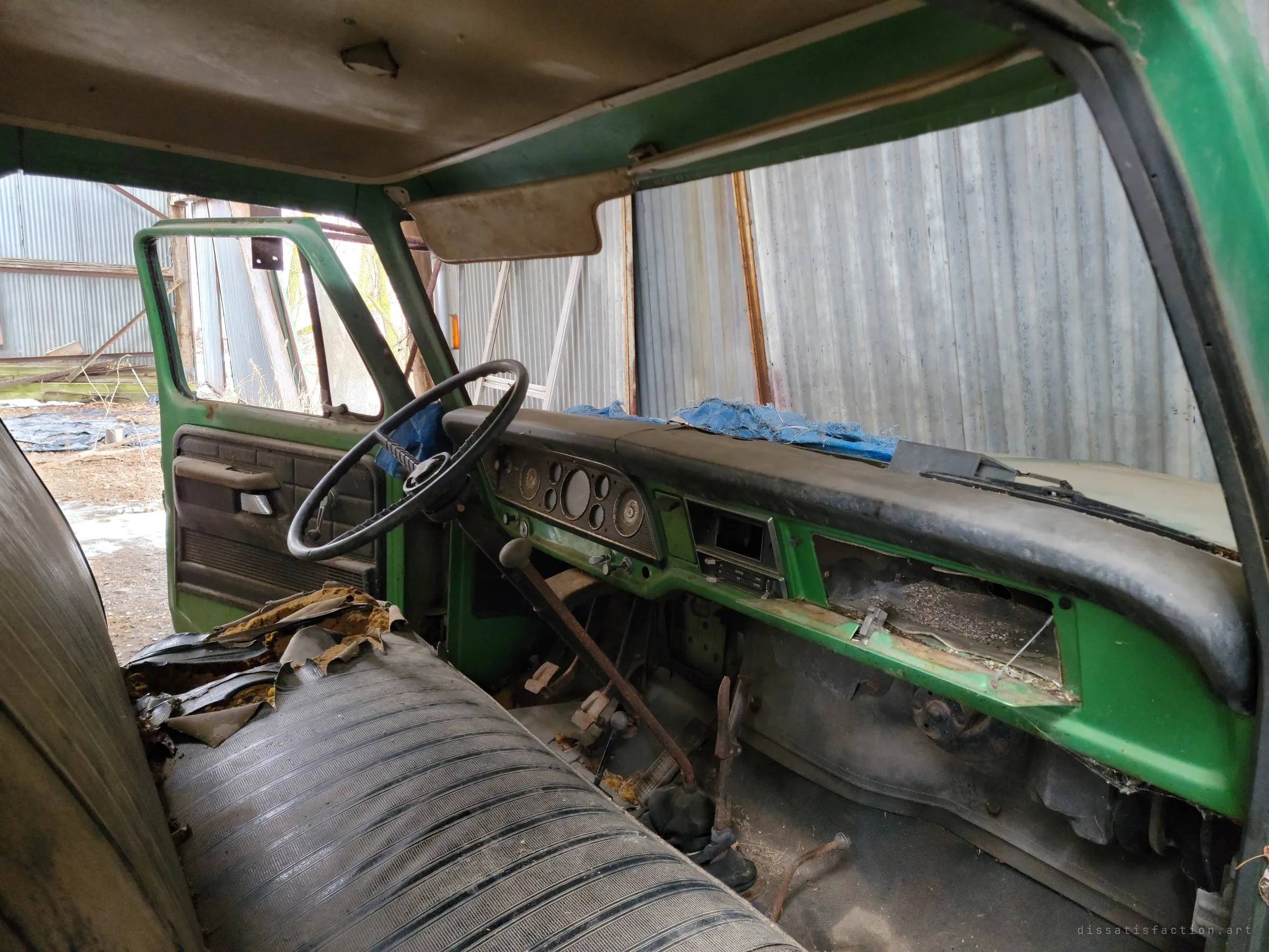 Inside the truck cab: a torn bench seat, a bare dashboard with an open glovebox and a frayed blue tarp on the dash. The corrugated shed wall fills the windshield.