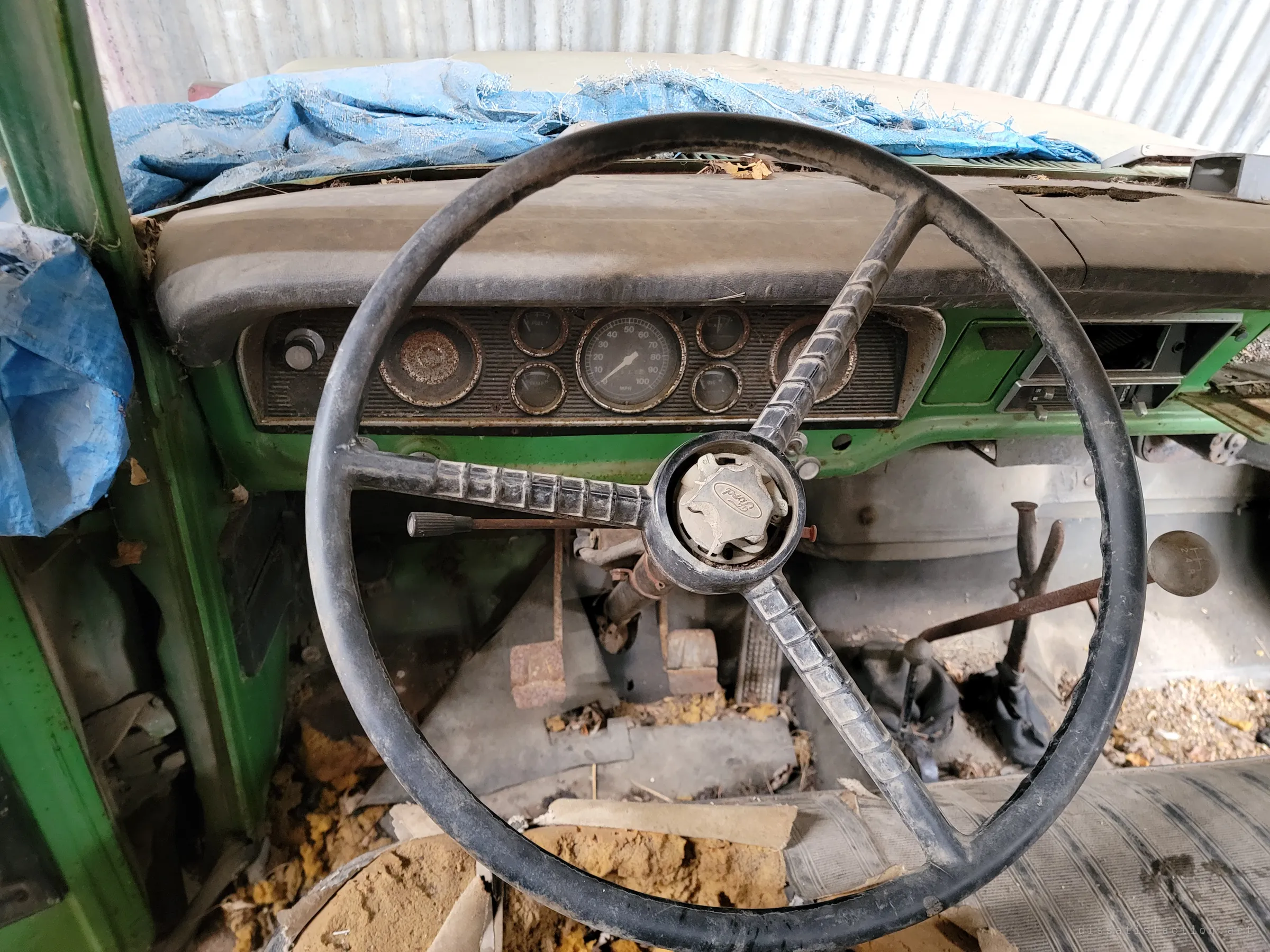 Close view over the thin black steering wheel to the truck's gauge cluster, rusted pedals and gear levers in a floor littered with foam and debris. Everything is coated in dust and decay.