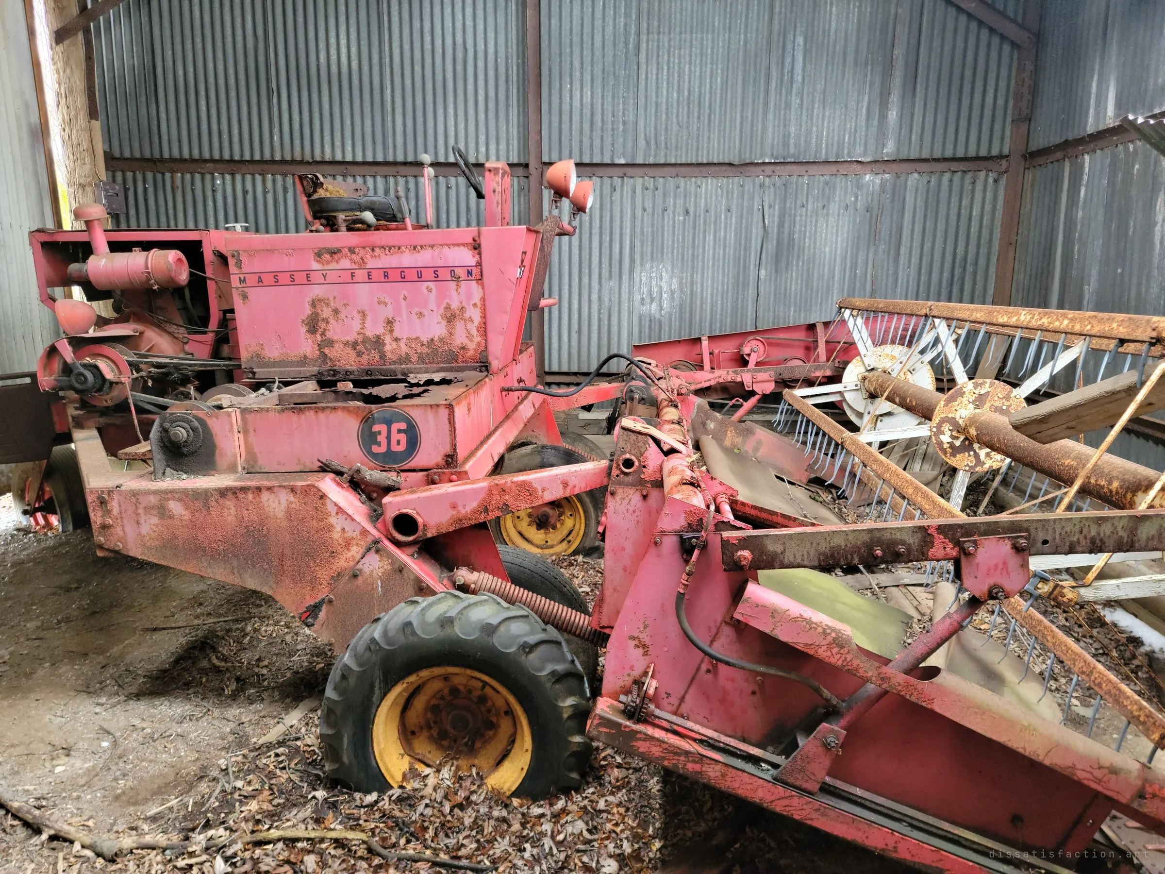 A faded red self-propelled combine with a rusted reel header, parked on a dirt floor inside a corrugated metal shed. The paint has worn through to rust across the machine.