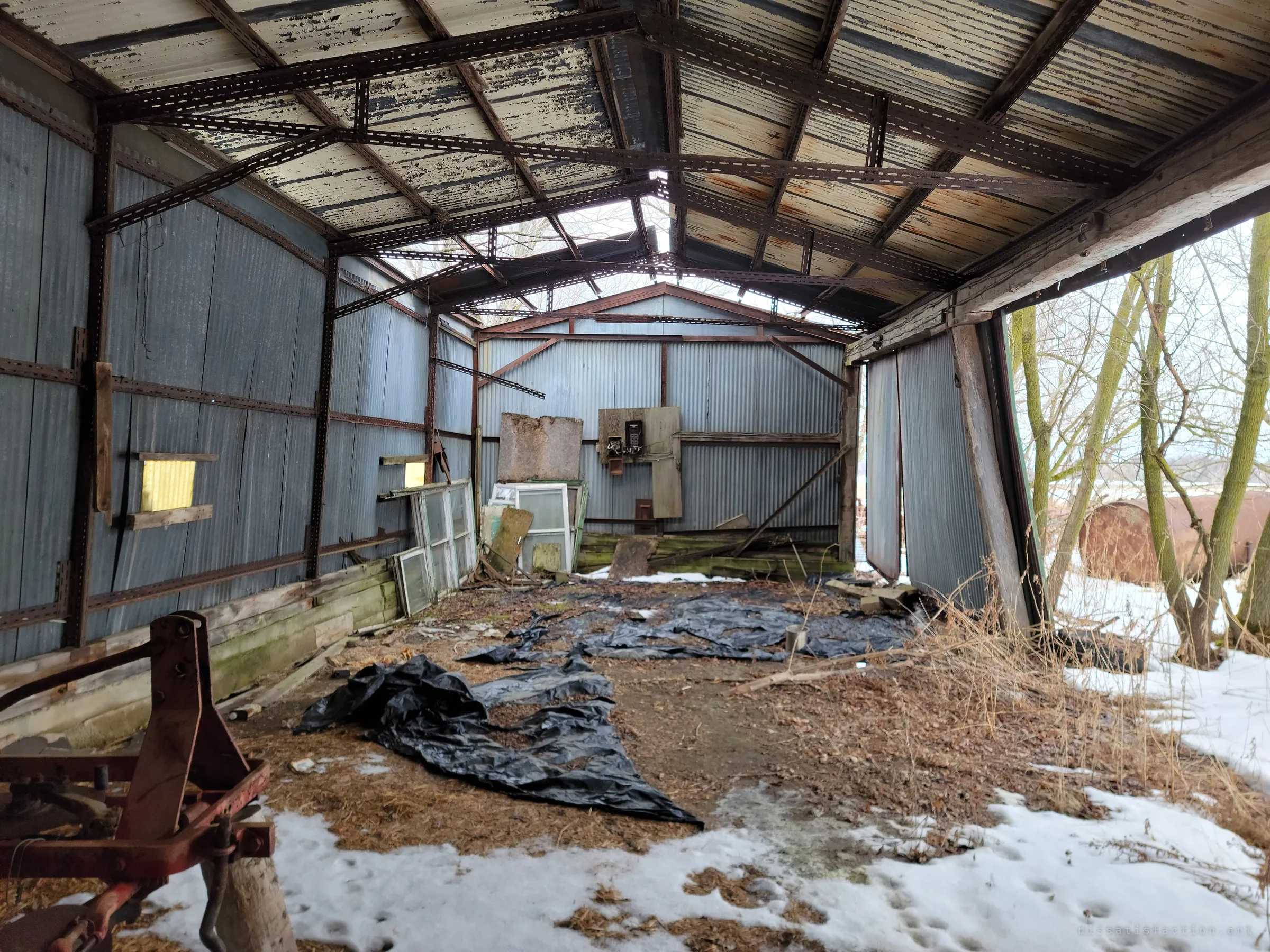 Inside an open-ended steel machine shed with a peeling corrugated roof and collapsing side panels, black plastic sheeting and old windows scattered over a snowy dirt floor. The rusted farm implement in the foreground and bare trees through the gap show how far the building has been let go.