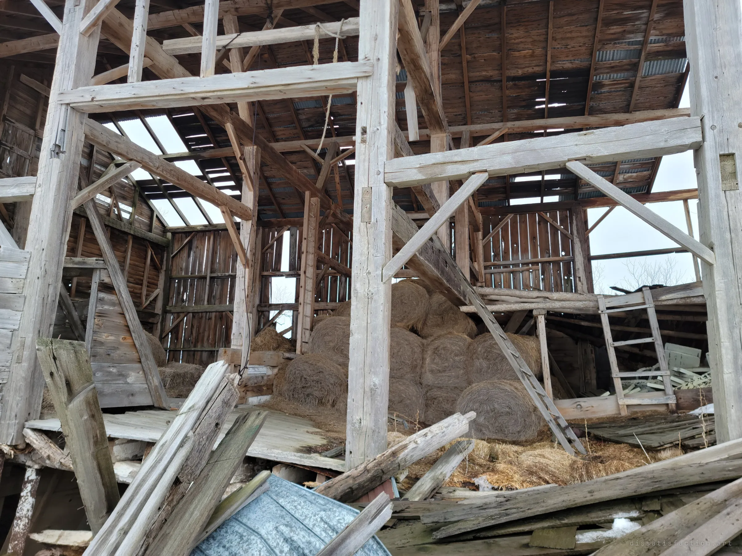 The hand-hewn timber frame of a barn open to the sky, round hay bales still stacked in the bay beneath a roof full of gaps, with a ladder leaning against the loft. Broken planks and bent roofing pile up in the foreground.