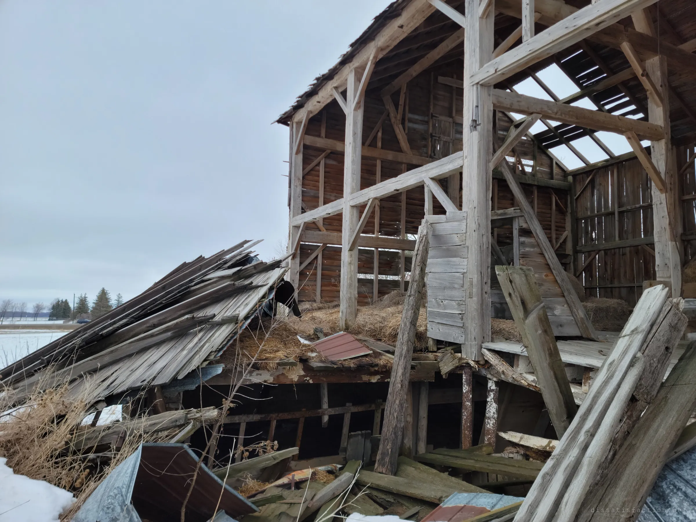 One side of the barn has caved in, its roof sliding down in a sheet of grey boards while the timber frame still stands open to an overcast sky. Snow-covered flat farmland stretches out beyond the collapse.