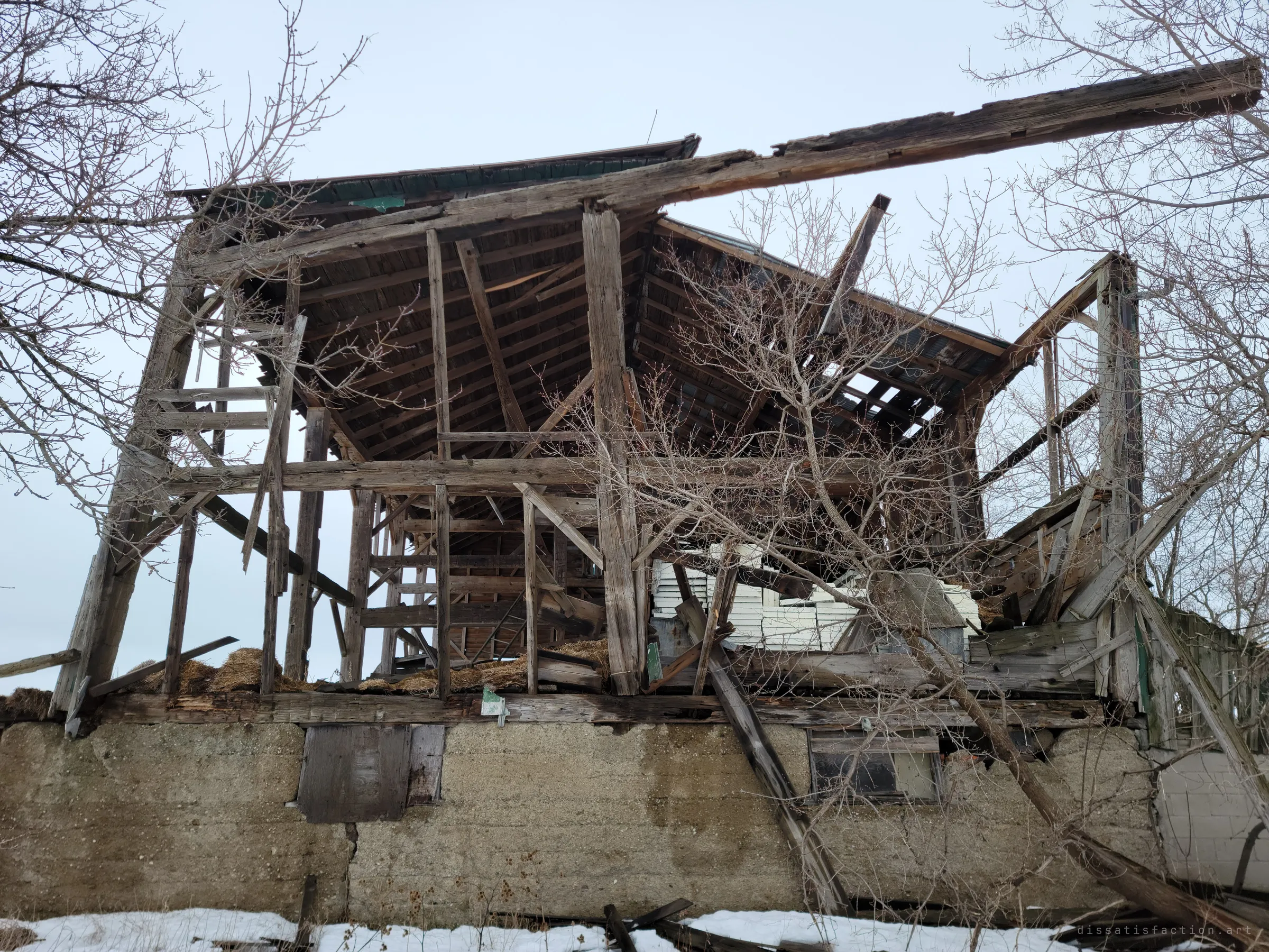Looking up at the barn from below its concrete foundation, the gutted timber frame and broken roof silhouetted against a white sky through bare branches. A long beam juts off the top like a snapped bone.