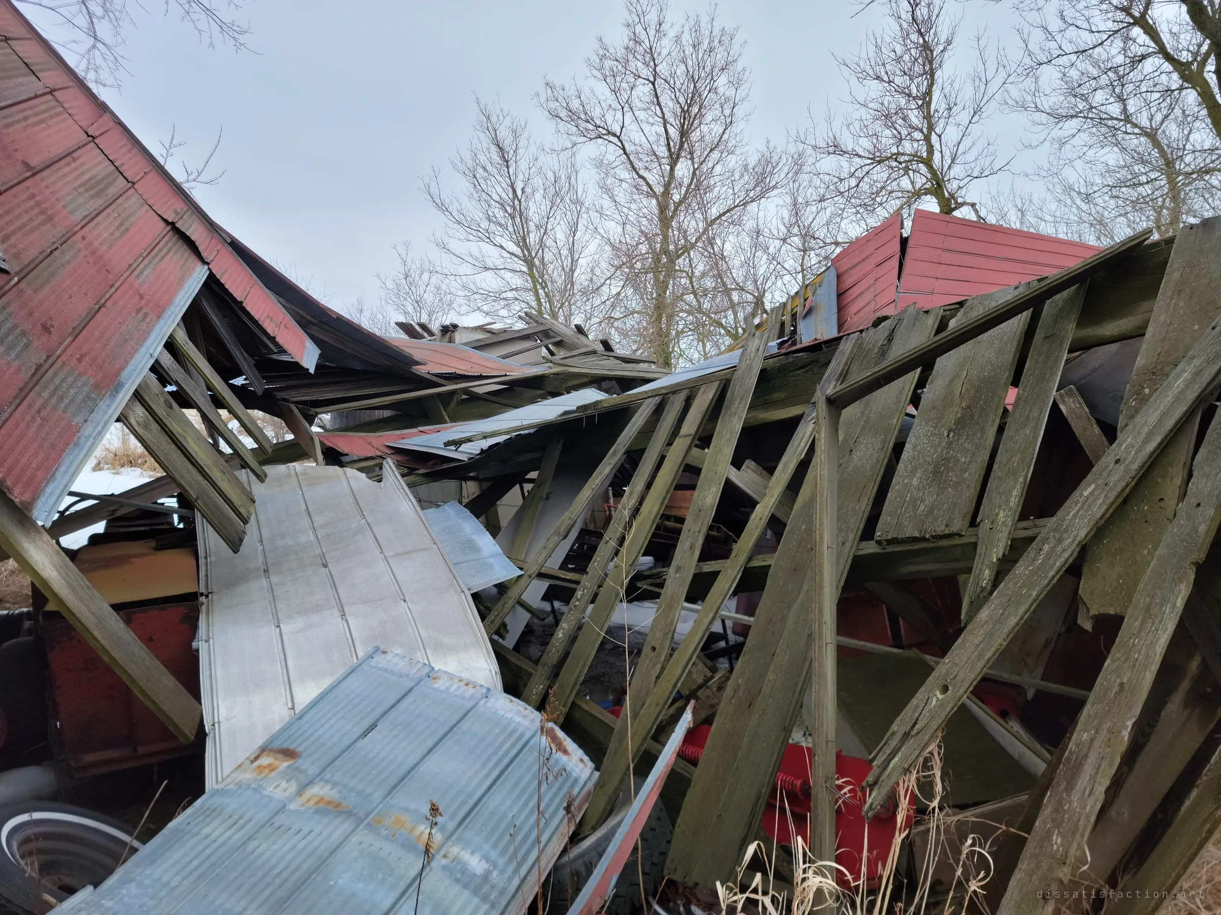 A heap of collapsed outbuilding: red metal siding folded over, grey boards leaning at every angle and old sheet-metal panels on top, with a whitewall tyre and red farm equipment buried underneath. The whole structure has come down on what it once sheltered.