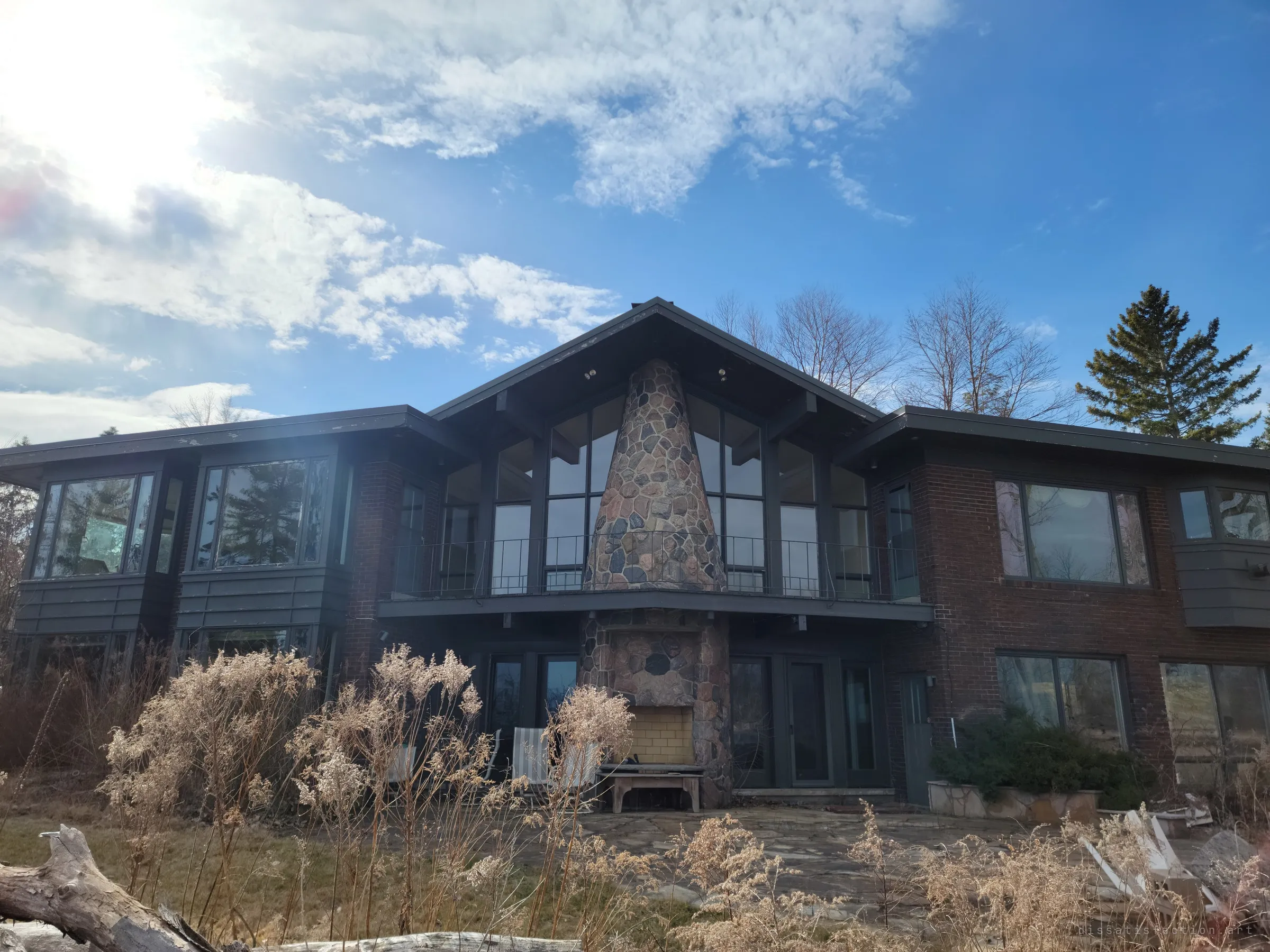 The lake side of a long mid-century house, a tapering fieldstone chimney splitting its glass gable, with dead goldenrod and driftwood across the overgrown lawn.