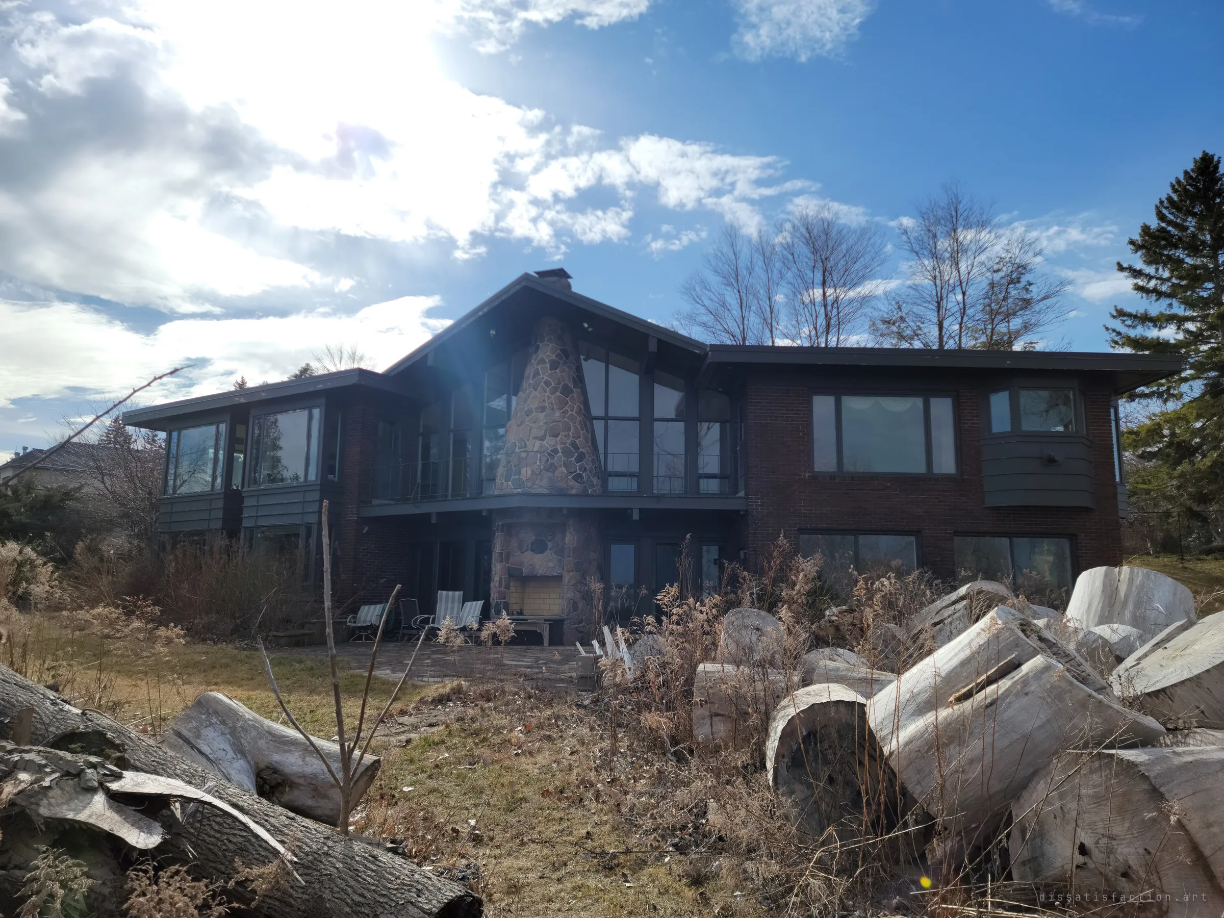 A wider view of the house from the yard, stacked pale logs in the foreground and patio chairs still set out around the outdoor fireplace.
