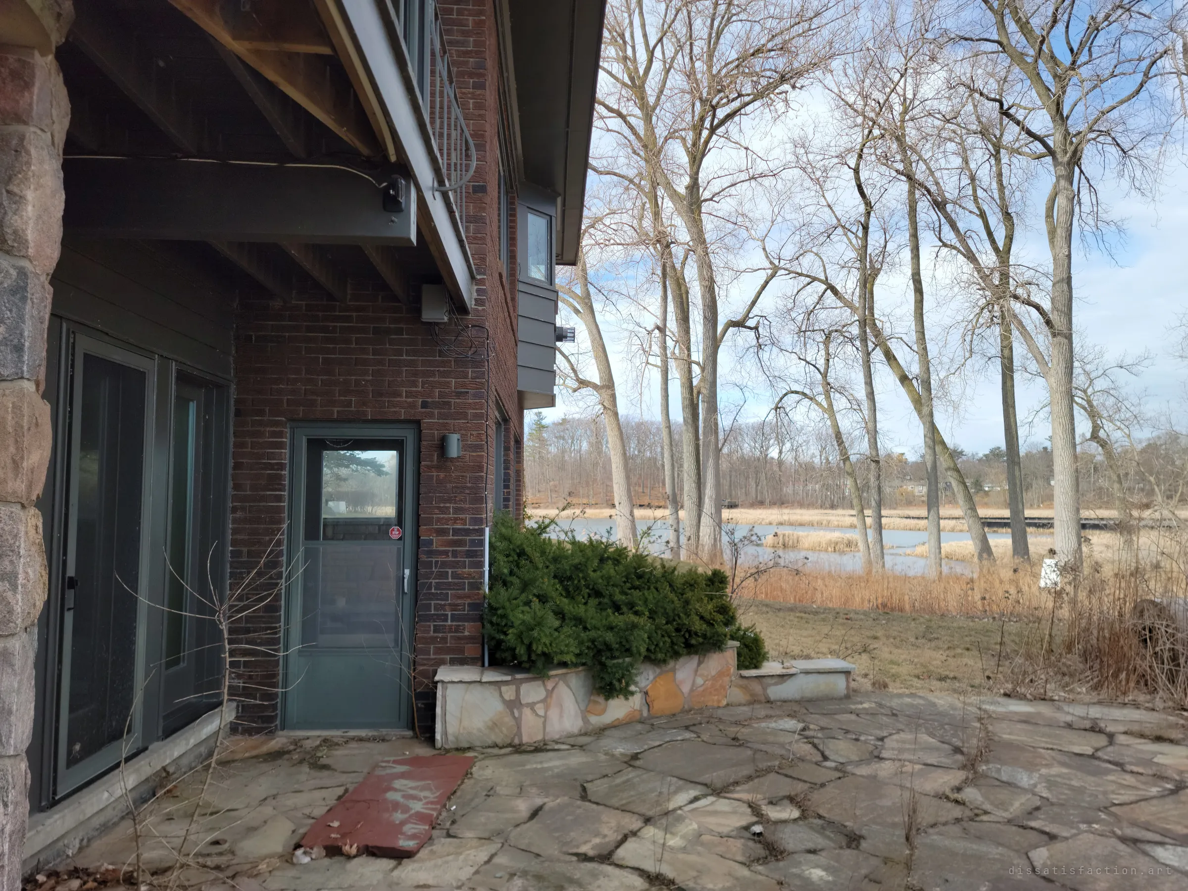 A side door under the cantilevered balcony opens onto a flagstone patio, with tall bare trees and a reedy pond beyond.
