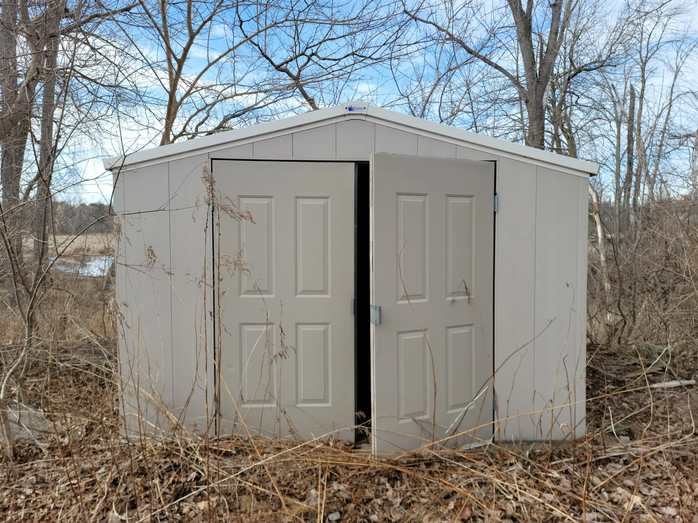 A plastic garden shed stands among bare saplings and dead grass, one door hanging open onto the dark inside.