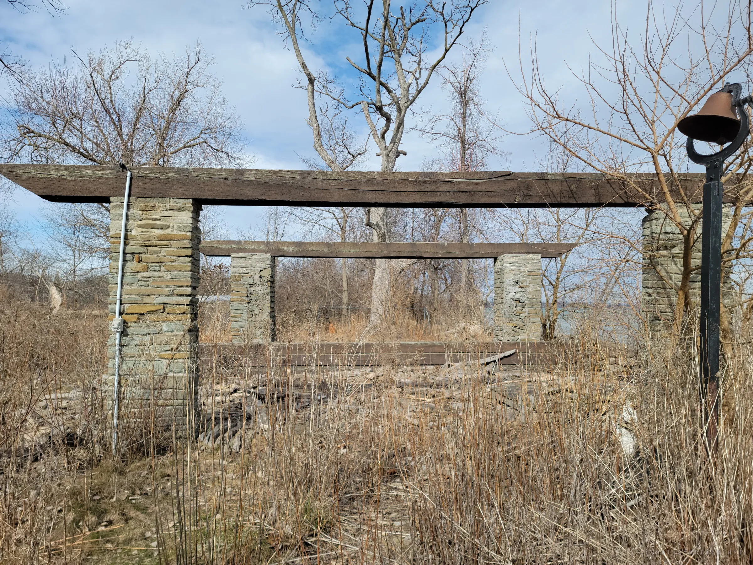 A stacked-stone pergola frame stands roofless in the tall dry grass near the water, an old iron dinner bell on a post at right.