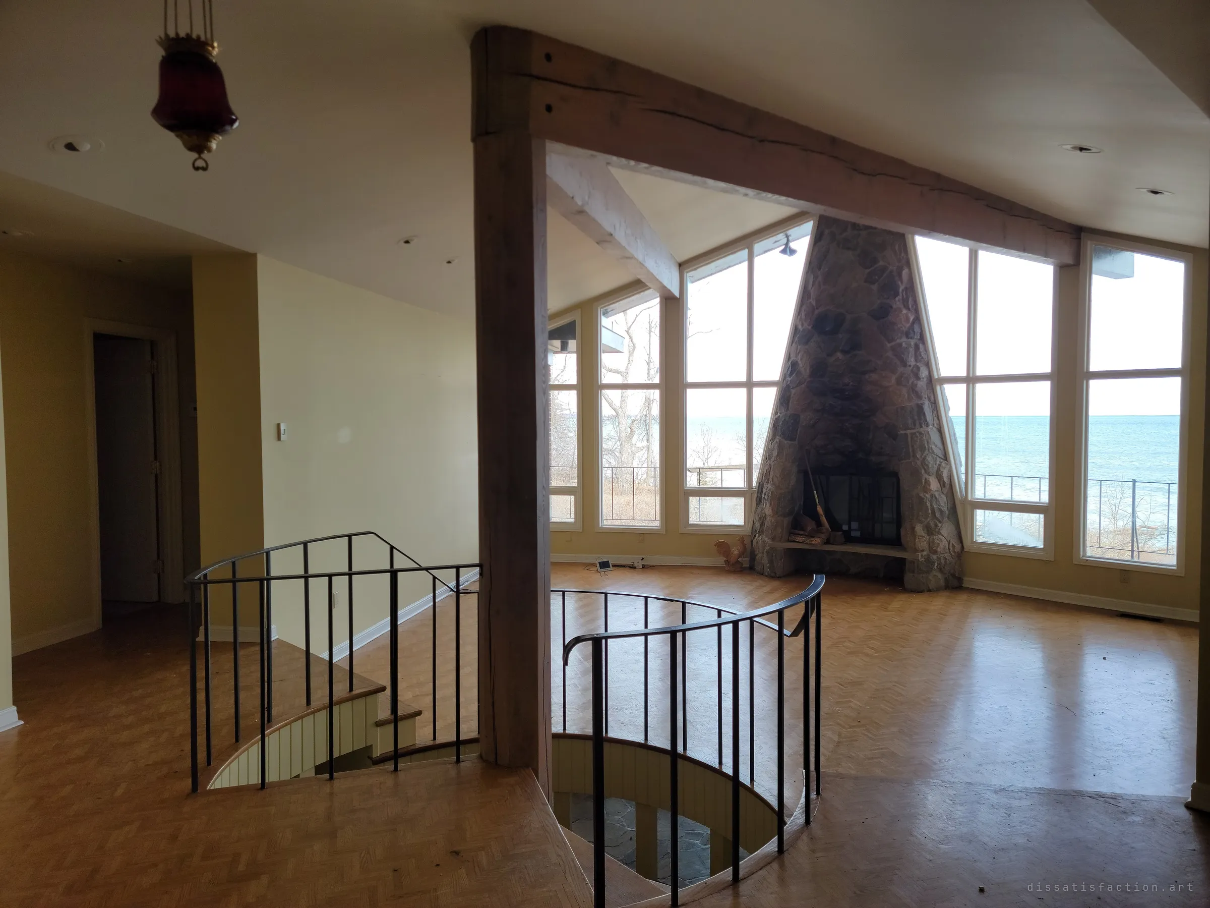 The empty great room, a round stair opening railed in black iron in front of a tapering stone fireplace set into a wall of windows onto the lake.