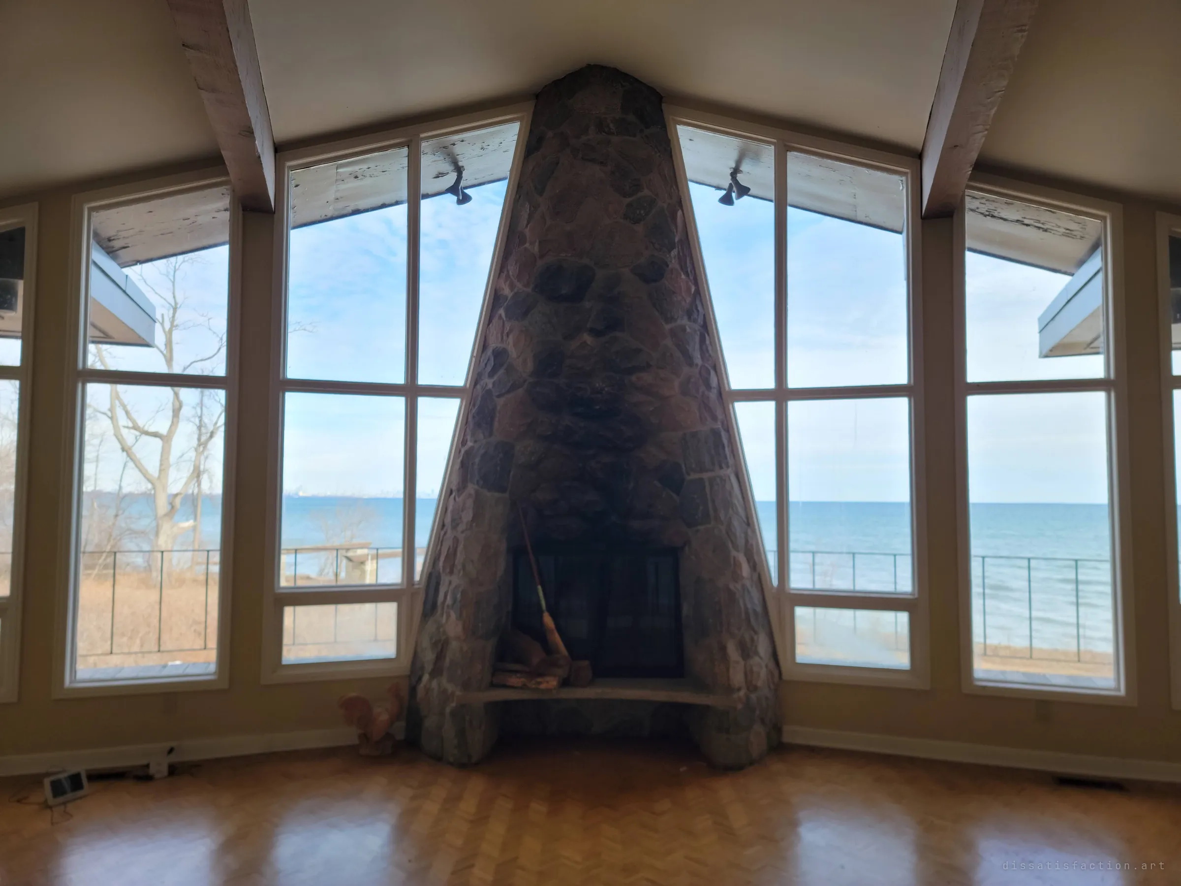 The stone fireplace head-on, rising to the peak of the ceiling between tall windows full of pale sky and lake.