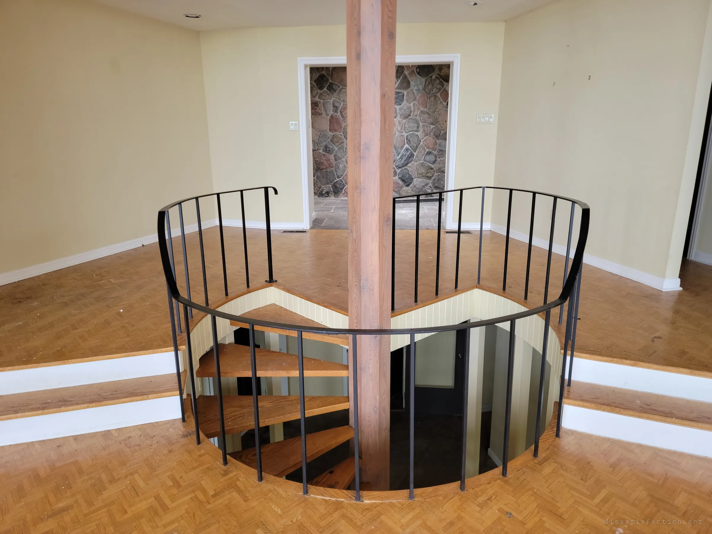 A spiral stair winds down around a timber post through a round opening in the parquet floor, a fieldstone wall glimpsed through the doorway behind.