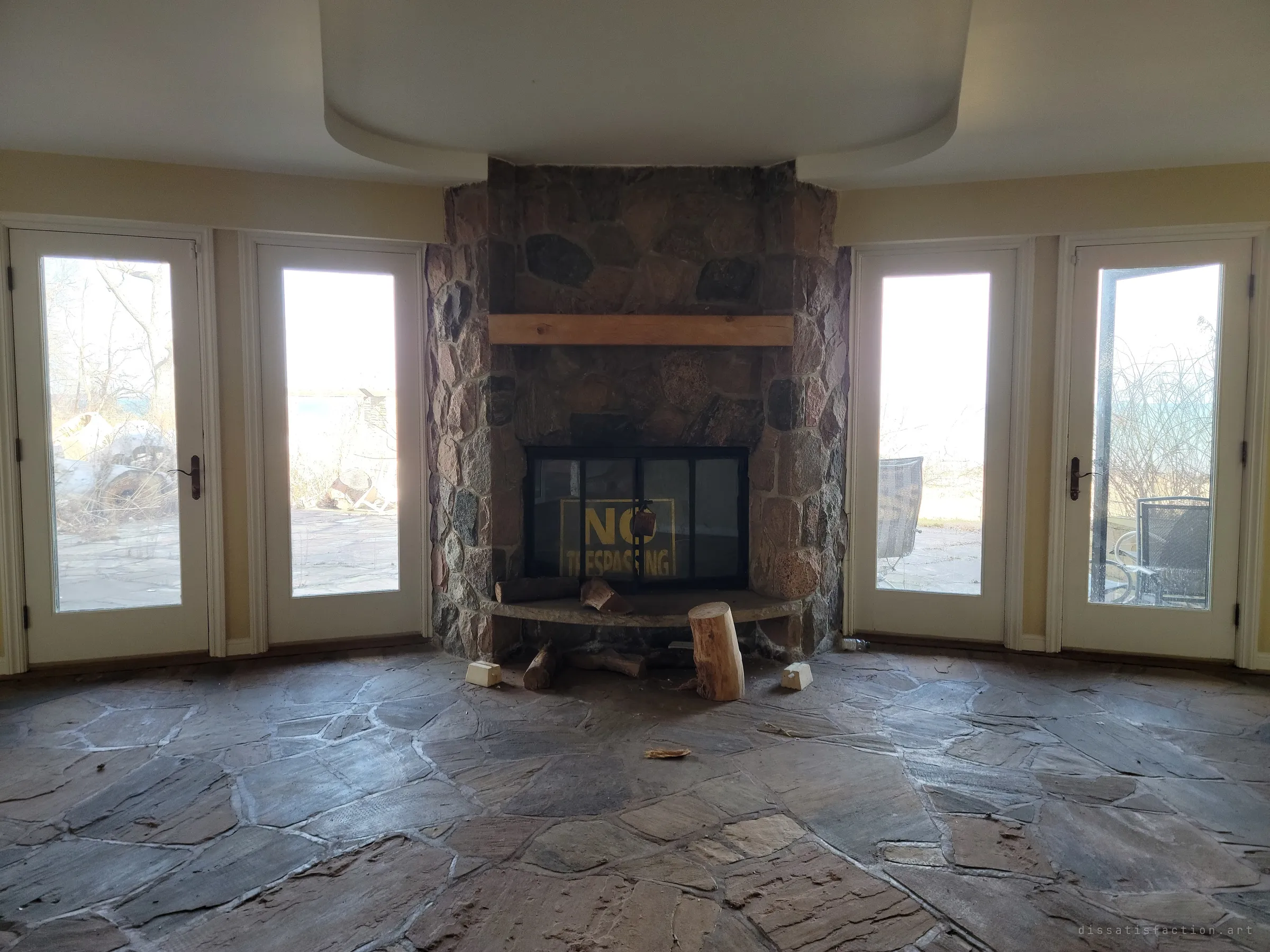 A lower-level room with a flagstone floor and a fieldstone fireplace between glass doors, a no-trespassing sign propped behind the fire screen.