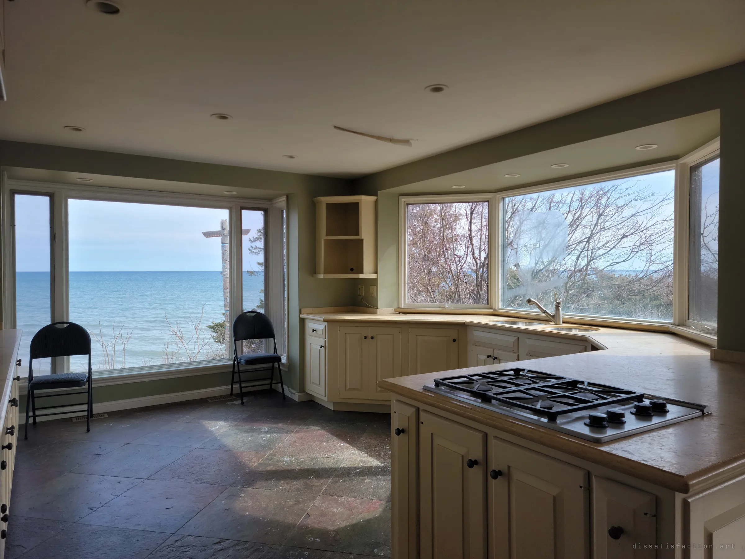 An abandoned kitchen with a gas cooktop island and bay windows, two folding chairs facing a wide window of open lake.