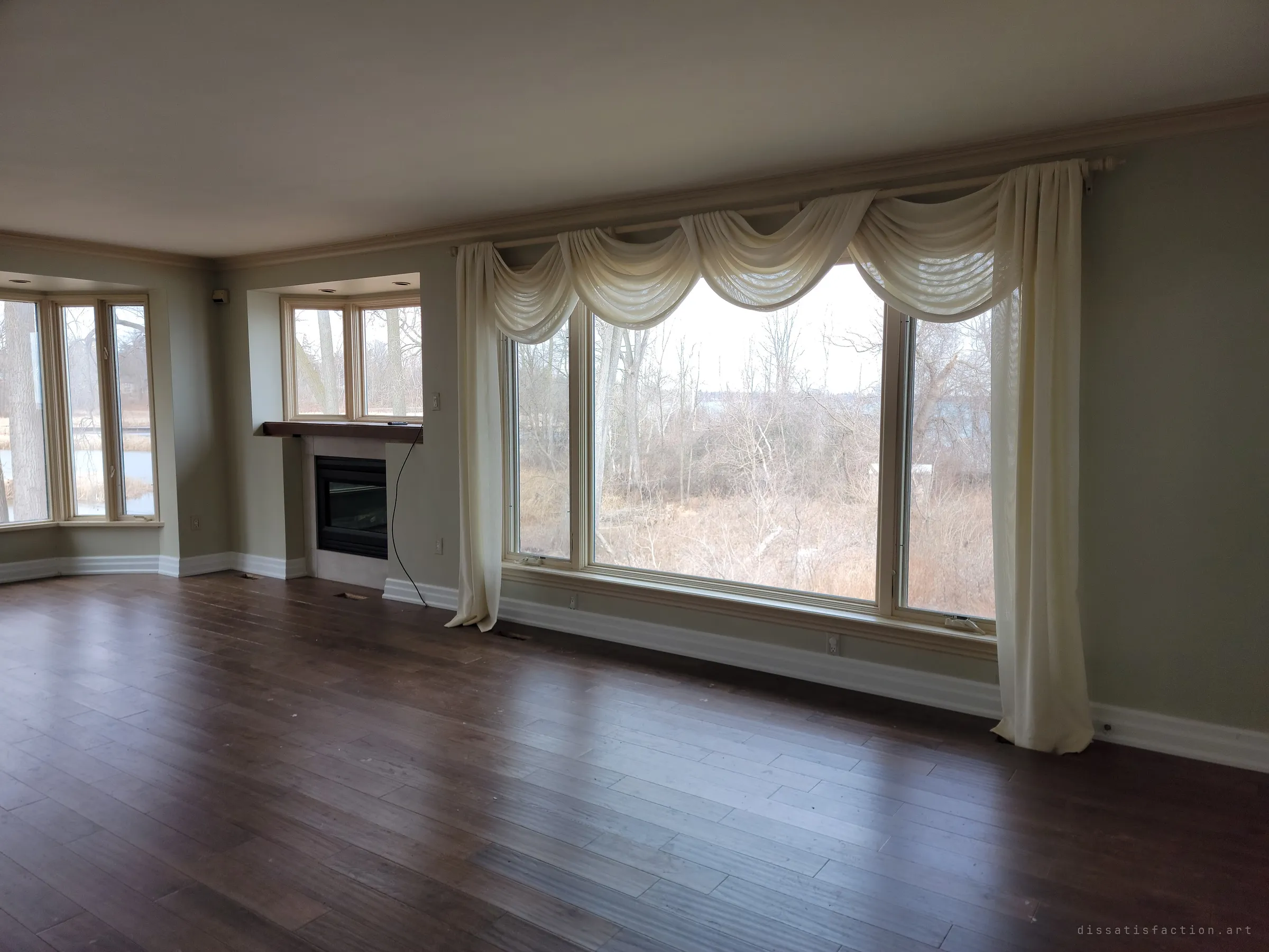 An empty living room with dark plank floors, a gas fireplace and a big window hung with sagging cream swags, looking out on bare winter brush. The house is cleared out, yet the drapes are still up.