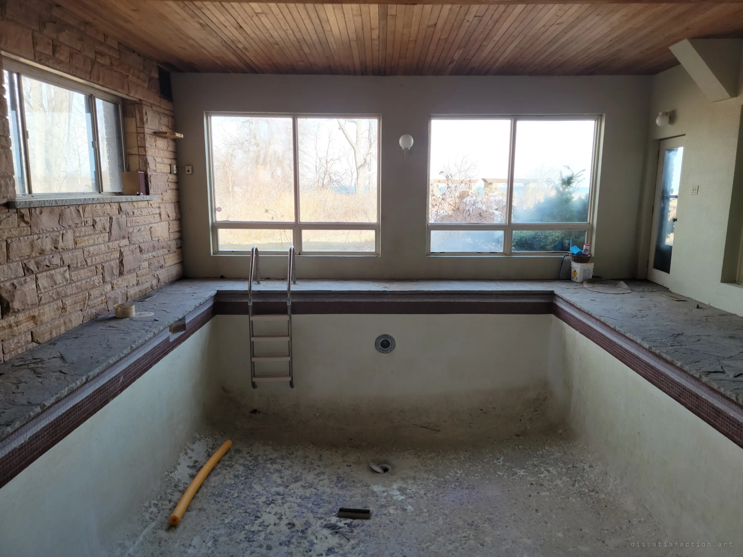 A drained indoor pool under a wooden ceiling, with its steel ladder still in place and a yellow pool noodle lying in the debris on the bottom. Stone walls and winter light through the windows make it feel like a room holding its breath.