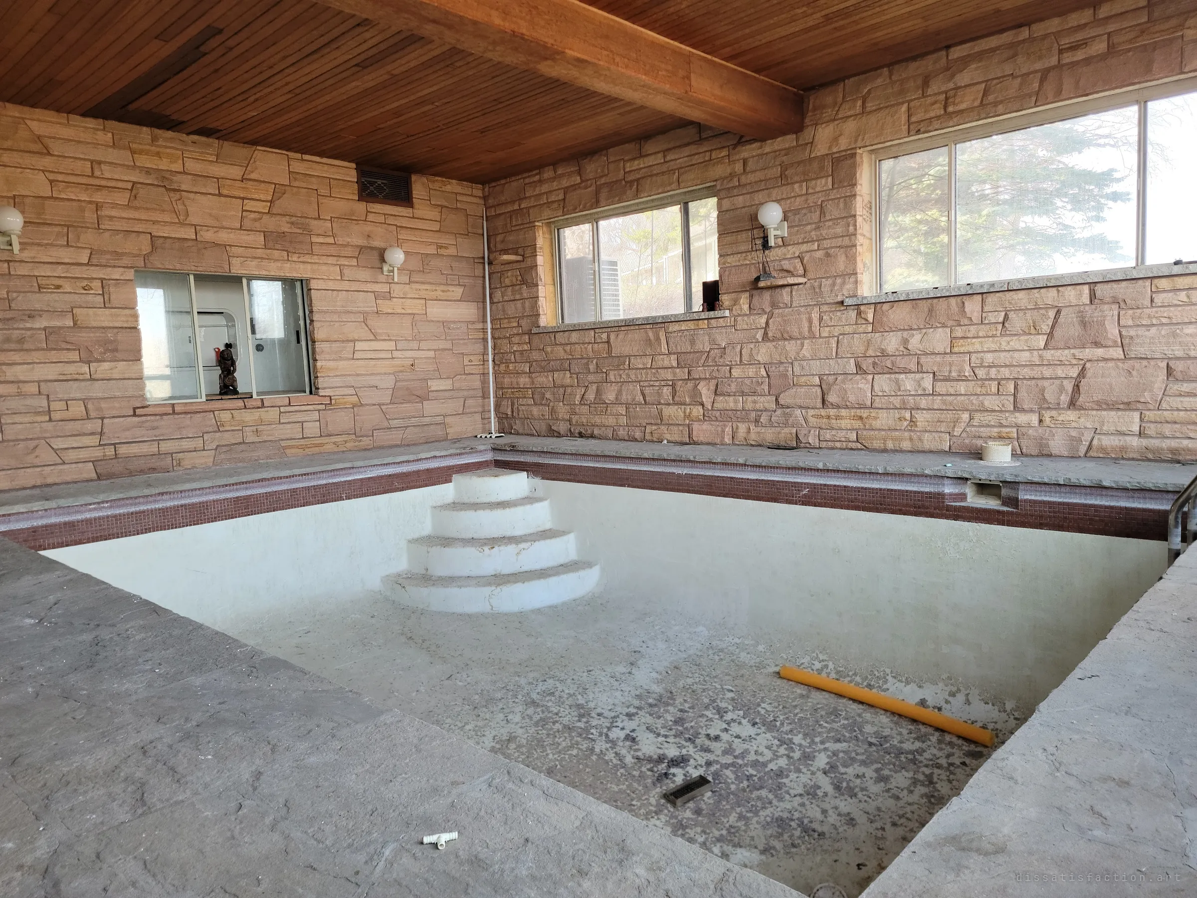 The same indoor pool from the shallow end: curved white steps lead down into the dry basin, framed by sandstone walls and globe lamps. A small figurine still stands in the pass-through window.