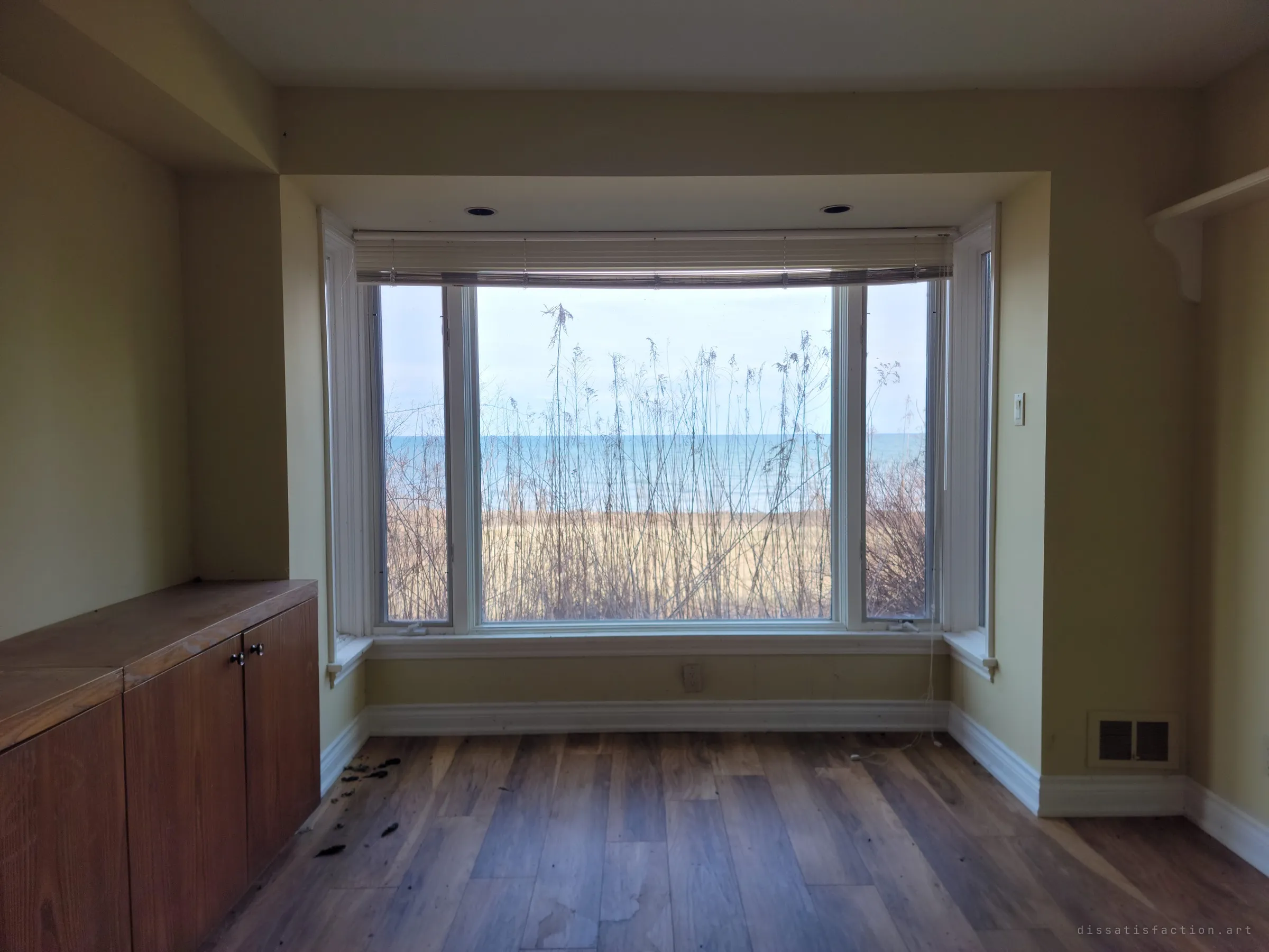 A pale bay window framed by dry reeds, opening straight onto a beach and flat blue water. The view the house was built for, now seen from an empty room.