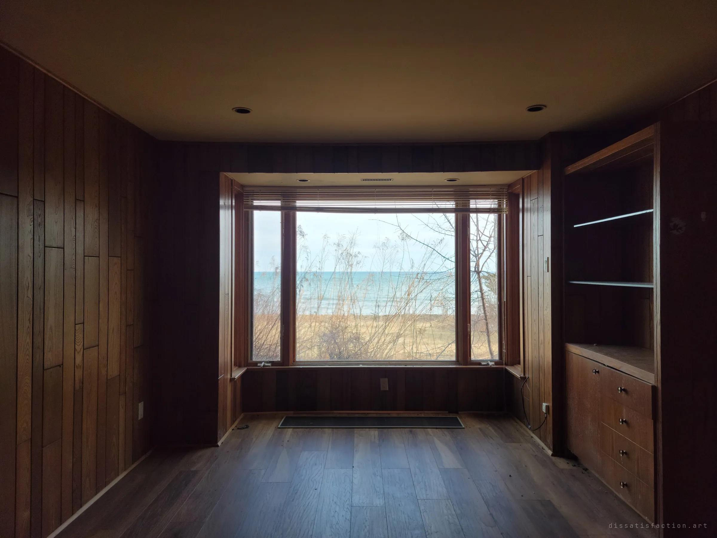 A dark wood-panelled den with built-in shelves, its bay window glowing with lake and sky through bare branches. The panelling swallows the light, so the window does all the work.