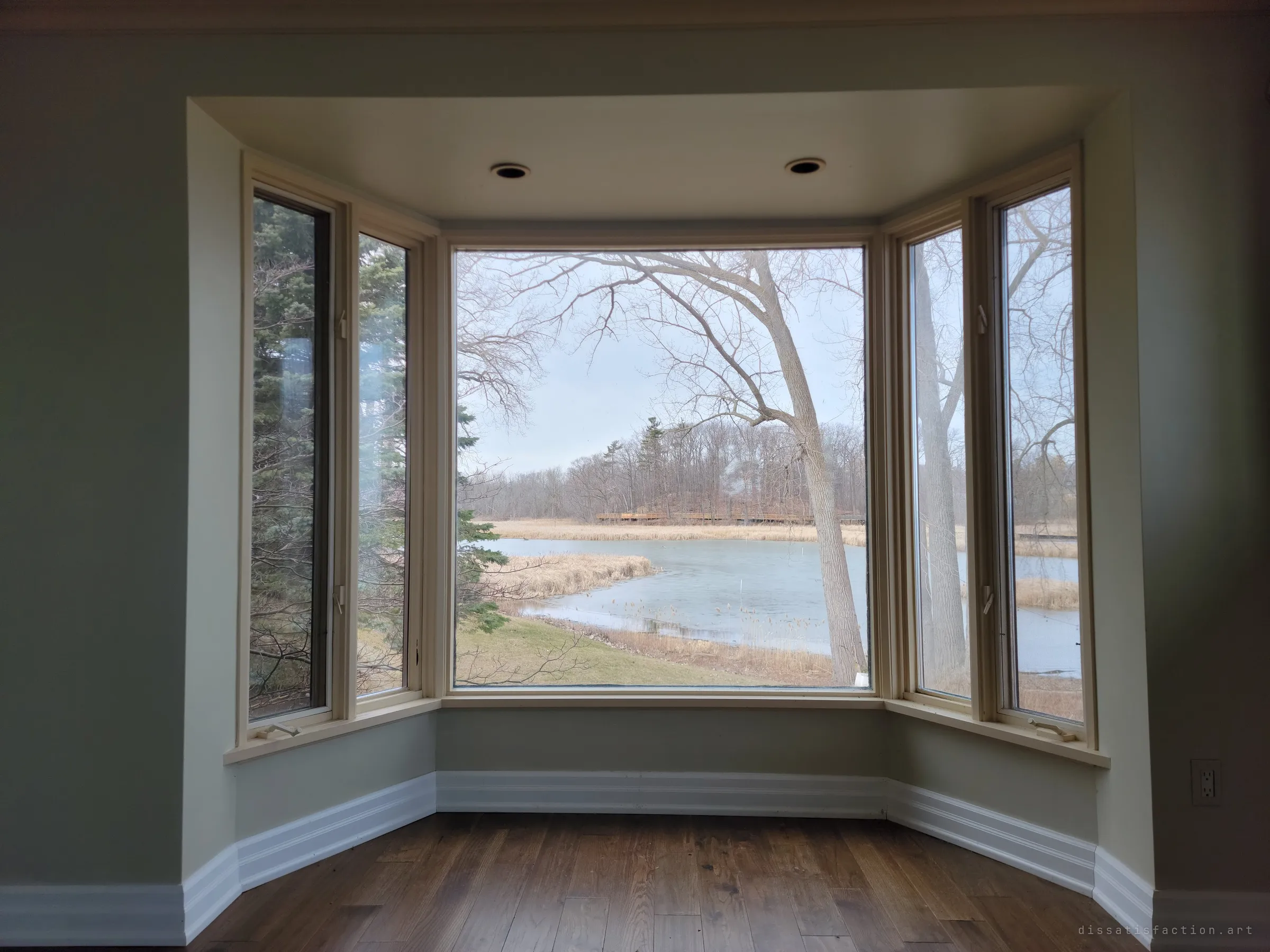 A bay window frames a half-frozen marsh pond, reeds and a bare tree leaning across the glass. Quiet and grey, the house turned its back on the lake here.
