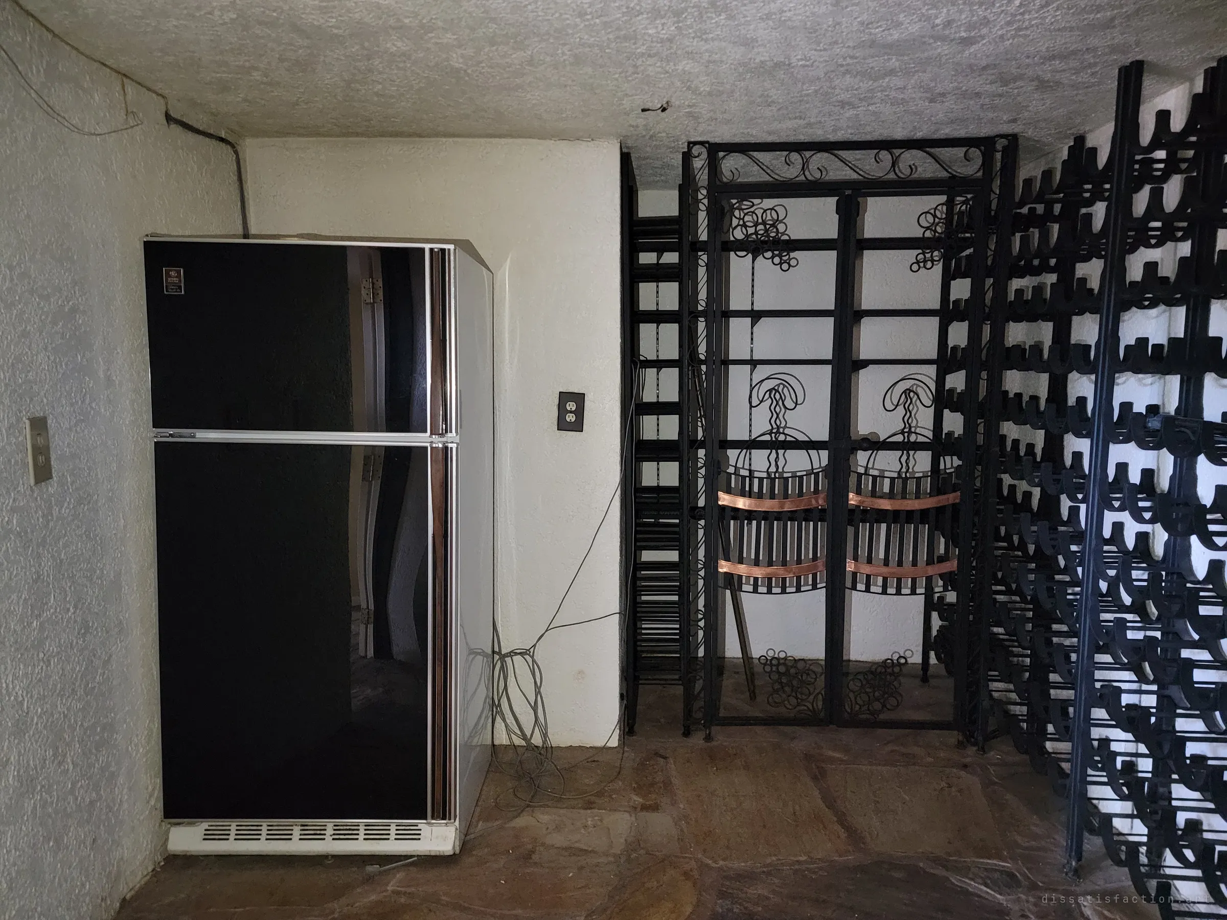 A basement wine cellar with a black-glass vintage fridge beside rows of wrought-iron bottle racks and a decorative gate with copper bands. Every rack is empty.