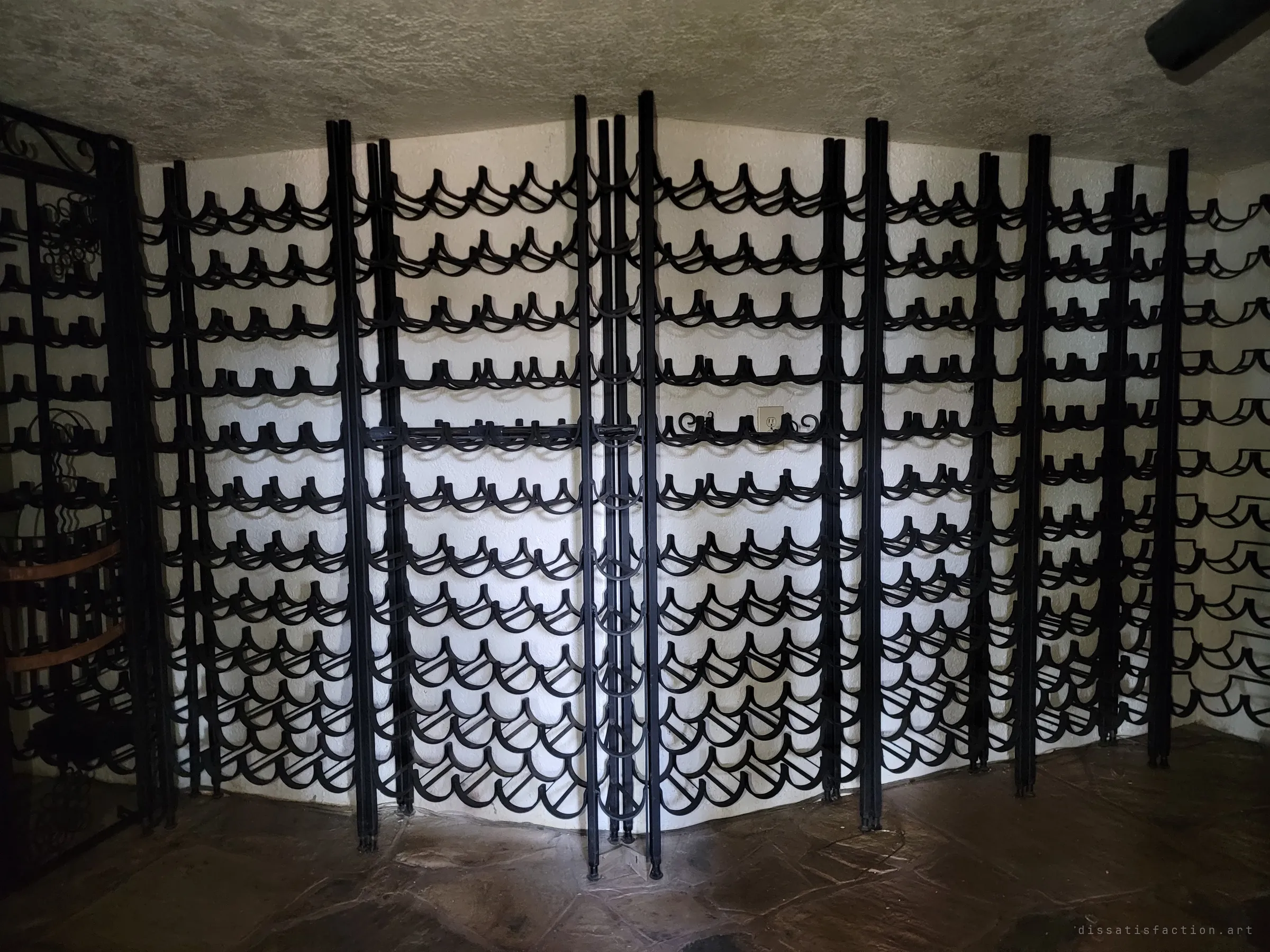 A wall of black wrought-iron wine racks, every cradle empty, lit hard by a flashlight against a white stucco cellar. The collection is long gone; only the scaffolding for it remains.