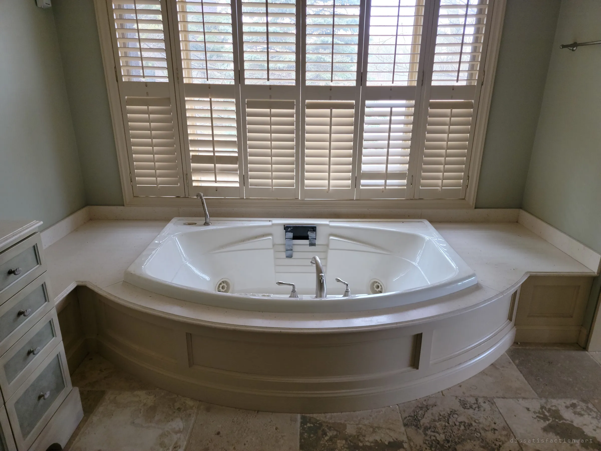 A deep jetted soaker tub set into a curved panelled surround beneath a wall of half-closed plantation shutters. Soft daylight through the slats makes the empty luxury bathroom feel quietly abandoned.