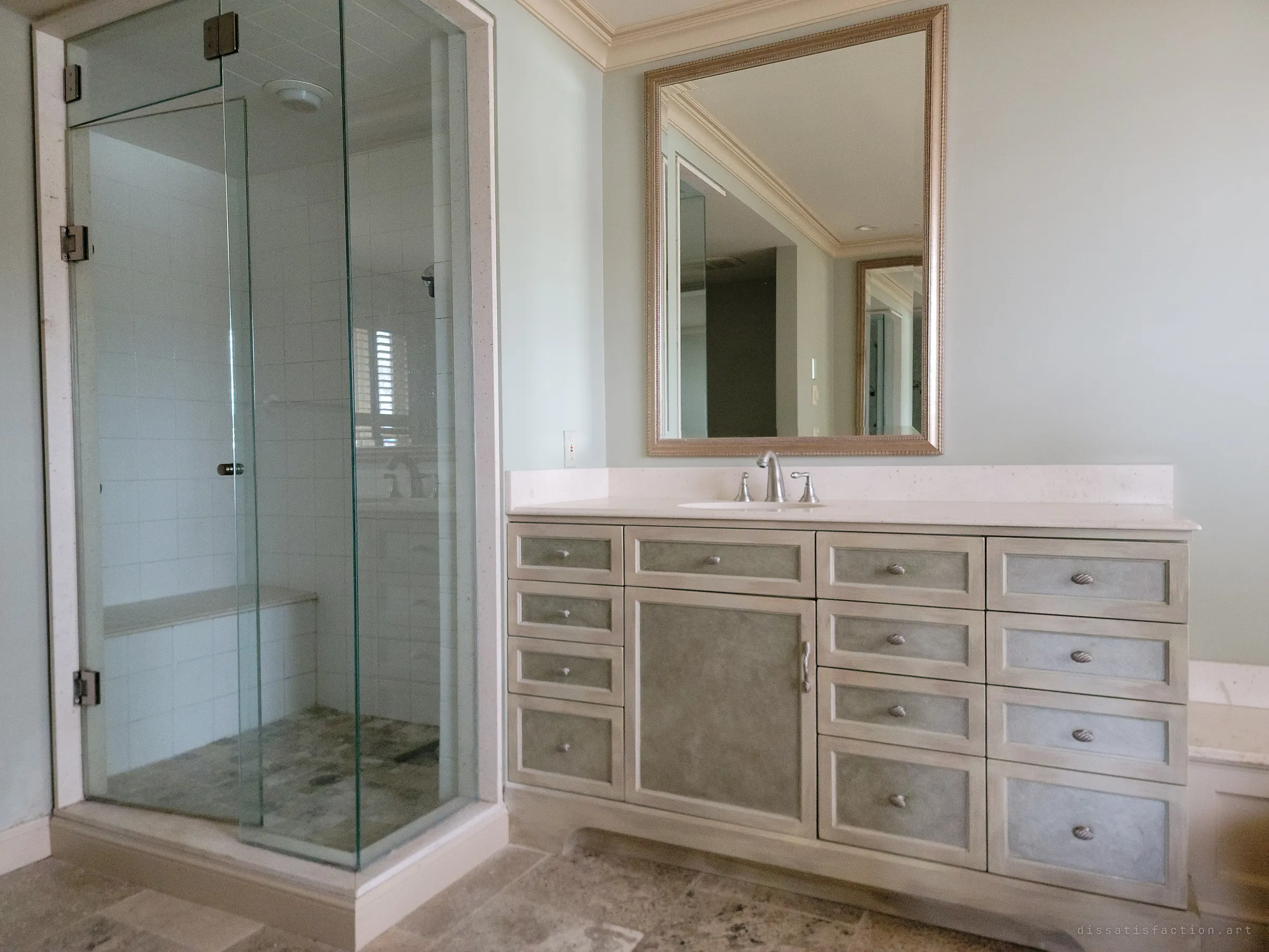 A glass steam shower beside a long painted vanity with a gilded mirror, the pale bathroom still clean and intact. The mirror reflects the empty rooms beyond.