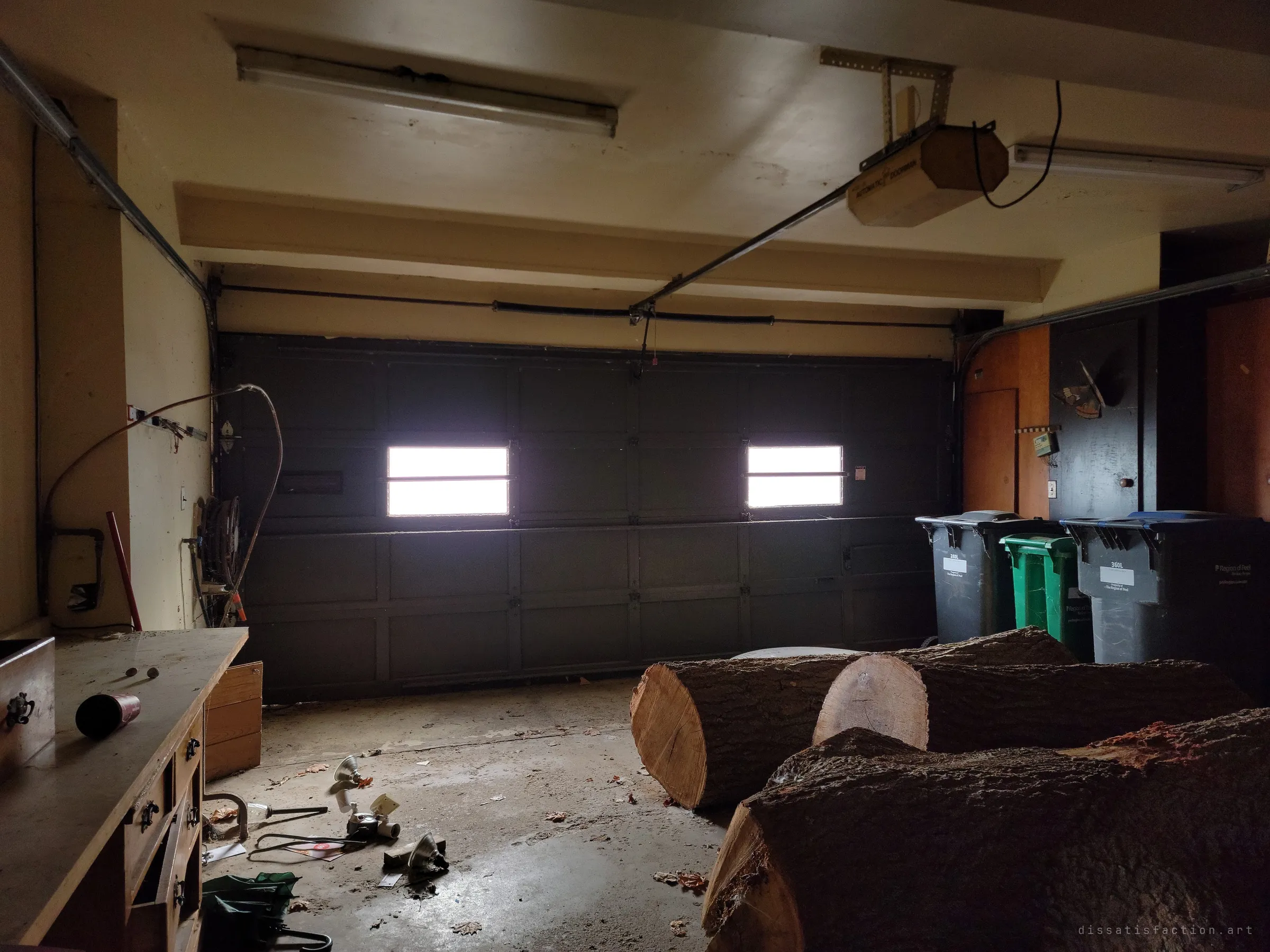 A dim two-car garage with light blazing through the small windows of the closed door, sawn tree trunks lying across the floor and old bins lined up against the wall. Scattered tools and debris show the house was left mid-task.