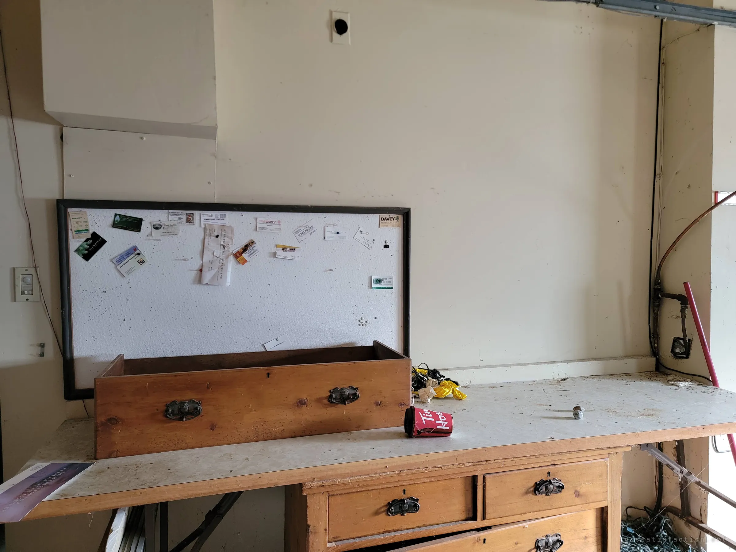 A workbench in the garage with an empty wooden drawer set in front of a corkboard still pinned with old business cards, a crushed coffee cup beside it. The house’s old routine of repairs and services is left on the wall.
