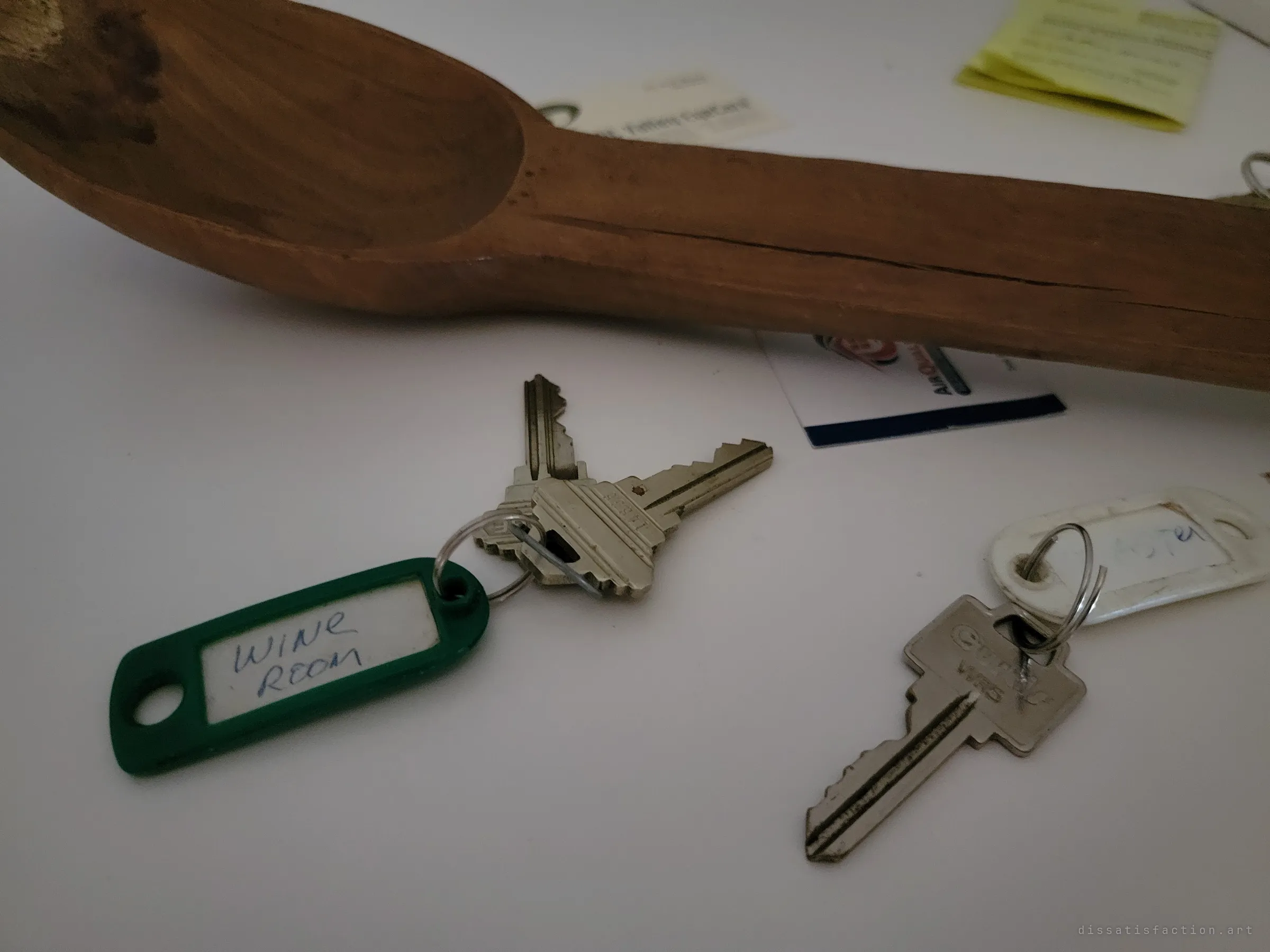Close-up of a key tagged by hand for the wine room lying under the carved wooden spoon, more keys and cards blurred behind. It ties the empty cellar back to the people who once locked it.